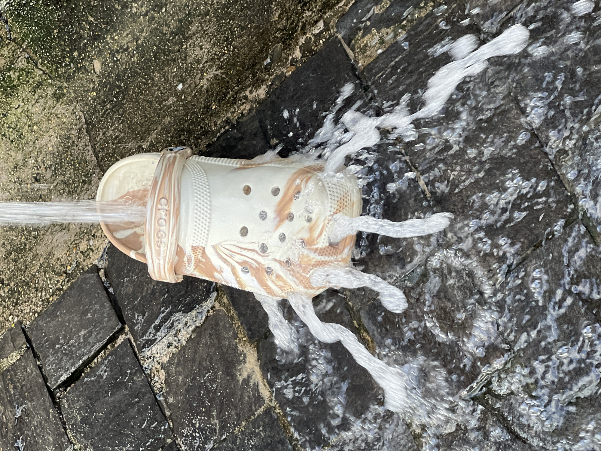 A cream-colored Croc shoe with a marbled brown and cream pattern is positioned on a wet, dark stone patio. Water is being poured from above into the shoe, causing it to overflow and create streams of foamy water that cascade down the sides and onto the patio. The patio is composed of irregular, dark gray stone tiles with some lighter colored gravel and moss in the cracks and surrounding areas. The lighting suggests it is daytime. The background shows more of the textured, greenish-brown concrete or stone ground. The image appears to be taken at ground level, looking down at the shoe and the surrounding wet surfaces. The scene is devoid of any people or animals.