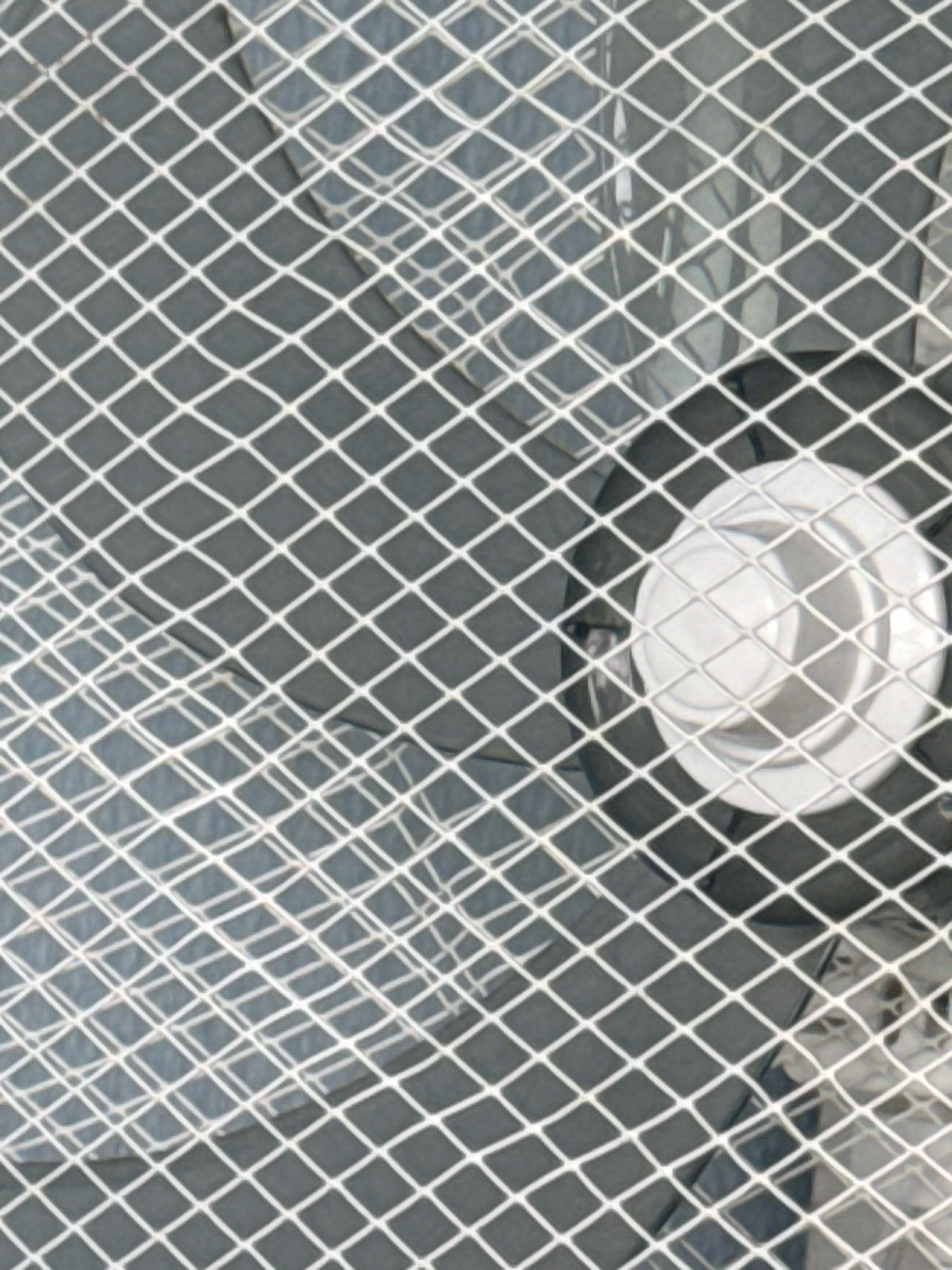 The image shows a close-up of a diamond-patterned metal grate in front of what appears to be the interior of a fan or similar electrical appliance. The fan has a white circular component with concentric rings in the center, and behind it, darker, blurred fan blades are visible. The grate has thin white lines forming diamond shapes. The background behind the grate is dark and textured, with some lighter, reflective areas suggesting a metallic surface. The lighting is even, and there are no people or animals in the frame. The scene is indoors.