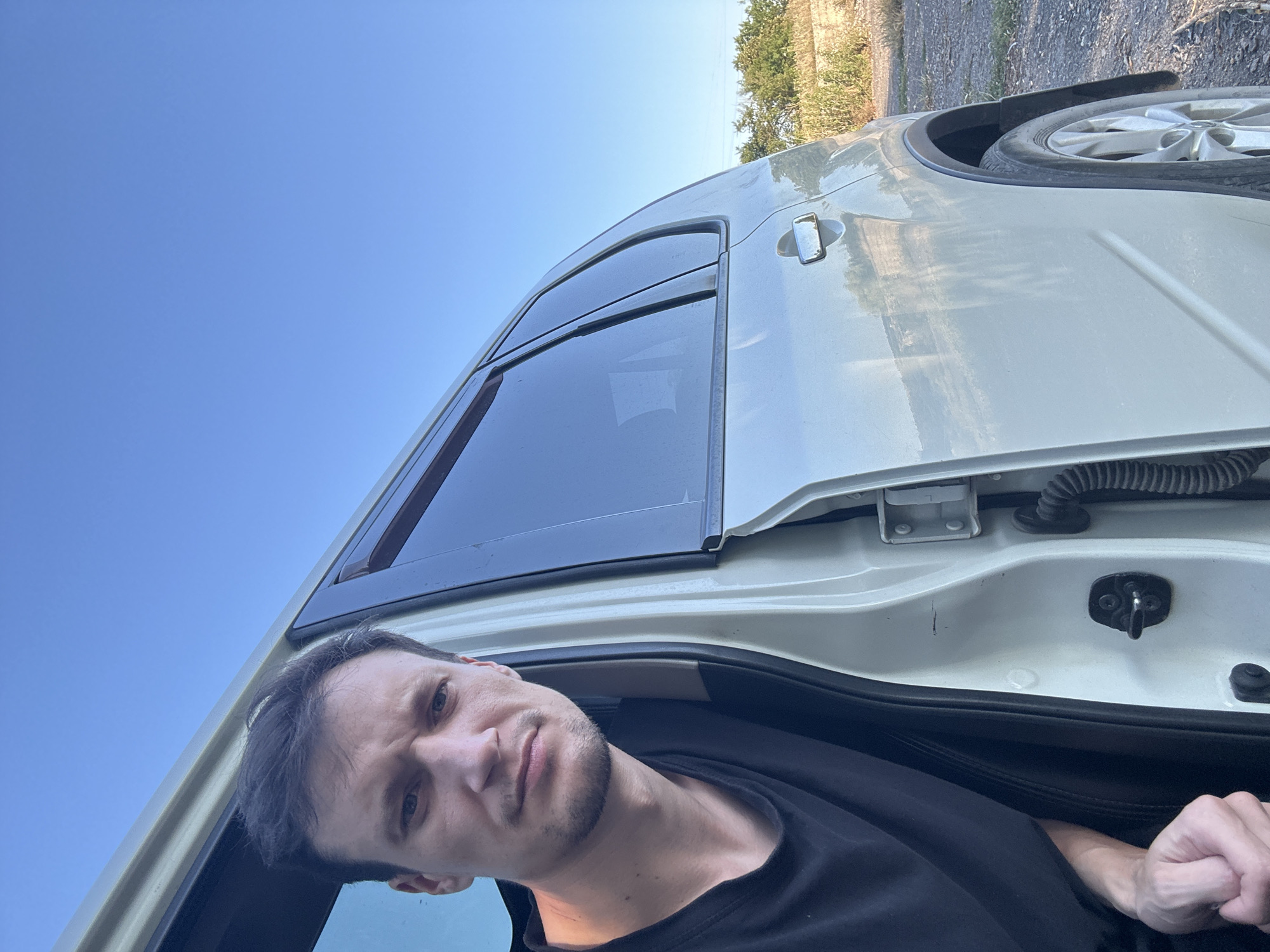 A man with short brown hair and stubble is looking into the camera from the open driver's side door of a light-colored SUV. He is wearing a black t-shirt. The car is parked on a dirt road next to dry grass and vegetation, with a clear blue sky overhead. In the background, there is a low wall and more vegetation. The image appears to have been taken in Makiv, Ukraine.