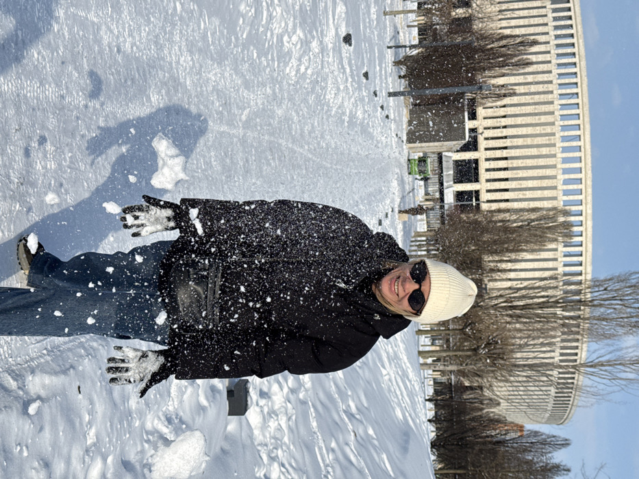 A female subject stands centrally in the foreground of a snow-covered outdoor scene, smiling directly at the viewer. She wears a white knit beanie, black sunglasses, a black puffer jacket, blue jeans, and white/grey gloves. A black fanny pack is visible across her torso. Snow particles are suspended in the air around her, adhering to her jacket and gloves, with a visible accumulation on her left glove and scattered particles around her, consistent with snow being thrown or disturbed. The ground is covered in deep, white snow, with a cleared, snow-dusted path leading into the background. Numerous bare deciduous trees are interspersed throughout the park-like environment. In the background, a large, modern, light-colored circular or oval stadium structure with vertical window slits is prominent, identifiable as the Krasnodar Stadium. A small green utility vehicle is discernible on the path further in the background. The sky is clear and blue, and strong sunlight casts distinct shadows on the snow. The scene is located in Plodorodny, Russia.