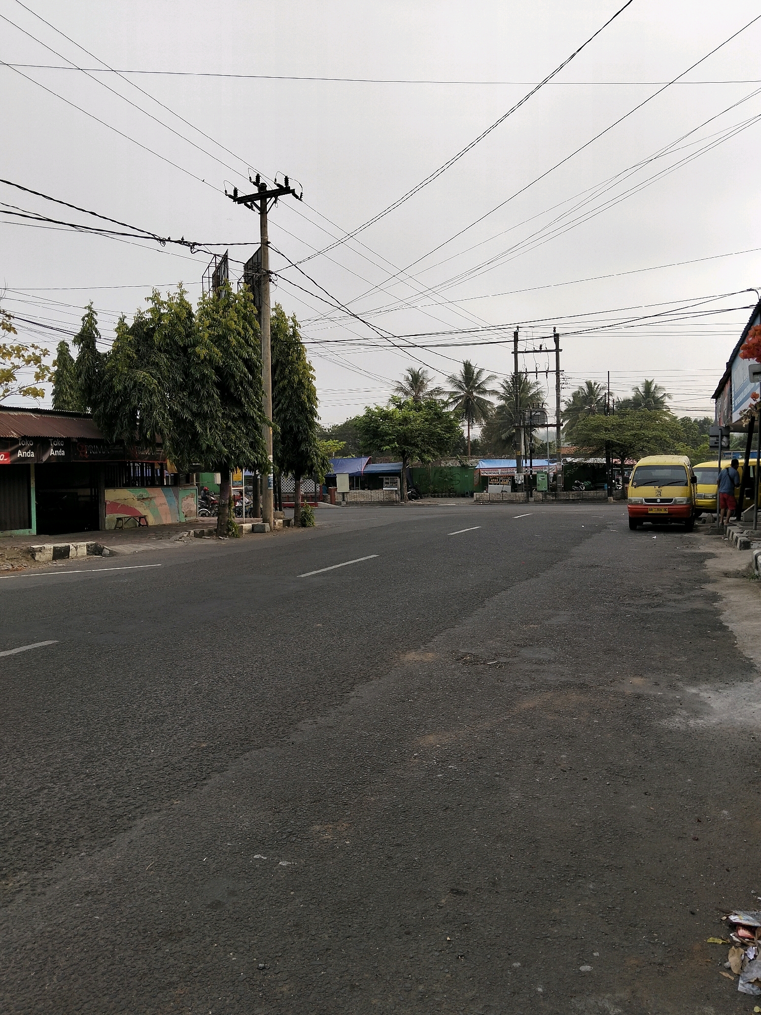 A cloudy day captures a street scene in Purworejo, Indonesia. The road, which appears to be asphalt with some water stains, curves to the right. On the left side of the road, a building with a corrugated metal roof and a mural painted on its wall is visible, with several large, leafy trees in front. Beyond the building, more trees and some structures with blue and green roofs can be seen. A utility pole with numerous wires crisscrosses the sky above. On the right side of the road, two yellow minibuses are parked. One of the minibuses has its license plate visible, reading "AA 1358 GC". Two men stand on the sidewalk next to the minibuses. Further down the road, more palm trees and utility poles are visible. Debris is scattered on the sidewalk and the edge of the road on the right.