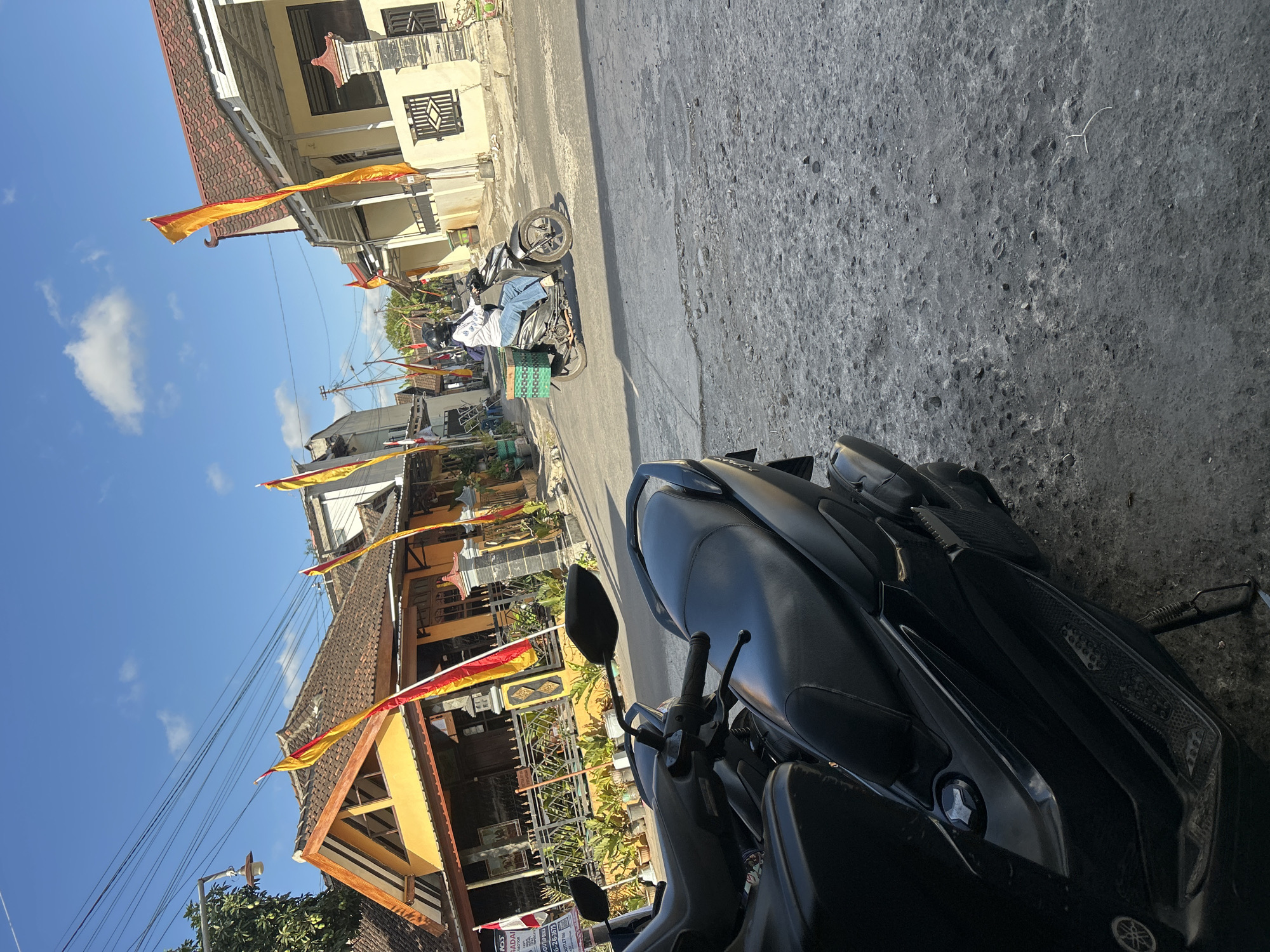 The image depicts a sunny street scene in Wonosari, Indonesia, with a clear blue sky and scattered clouds. In the foreground, a dark-colored scooter is parked on a textured gray pavement. Further down the street, a person wearing a white shirt, blue jeans, and a black helmet is riding another scooter. This scooter has a green woven basket attached to its rear. The street is lined with houses, one of which is painted yellow with a red-tiled roof. Red and yellow flags are prominently displayed along the street, attached to poles and fluttering in the breeze. The houses have fences and potted plants in front of them. Electrical wires are visible overhead. The overall impression is a quiet residential street on a bright day.