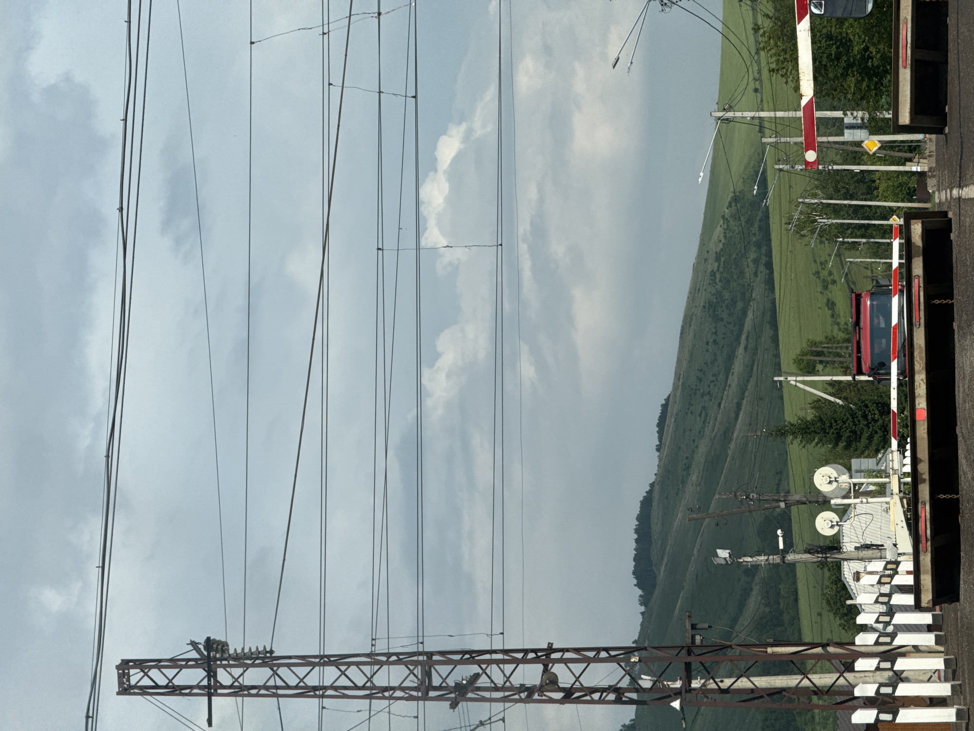 The image depicts a railway crossing in the city of Zykovo, Russia. The foreground shows a raised railway barrier, painted with red and white stripes, blocking a road. A red truck is visible beyond the barrier, waiting to cross the tracks. Overhead, a complex network of electrical wires crisscrosses the sky, supported by tall metal pylons and wooden poles. In the background, rolling green hills under a cloudy sky form the natural landscape. The overall scene suggests a typical day at a rural or semi-rural railway crossing.