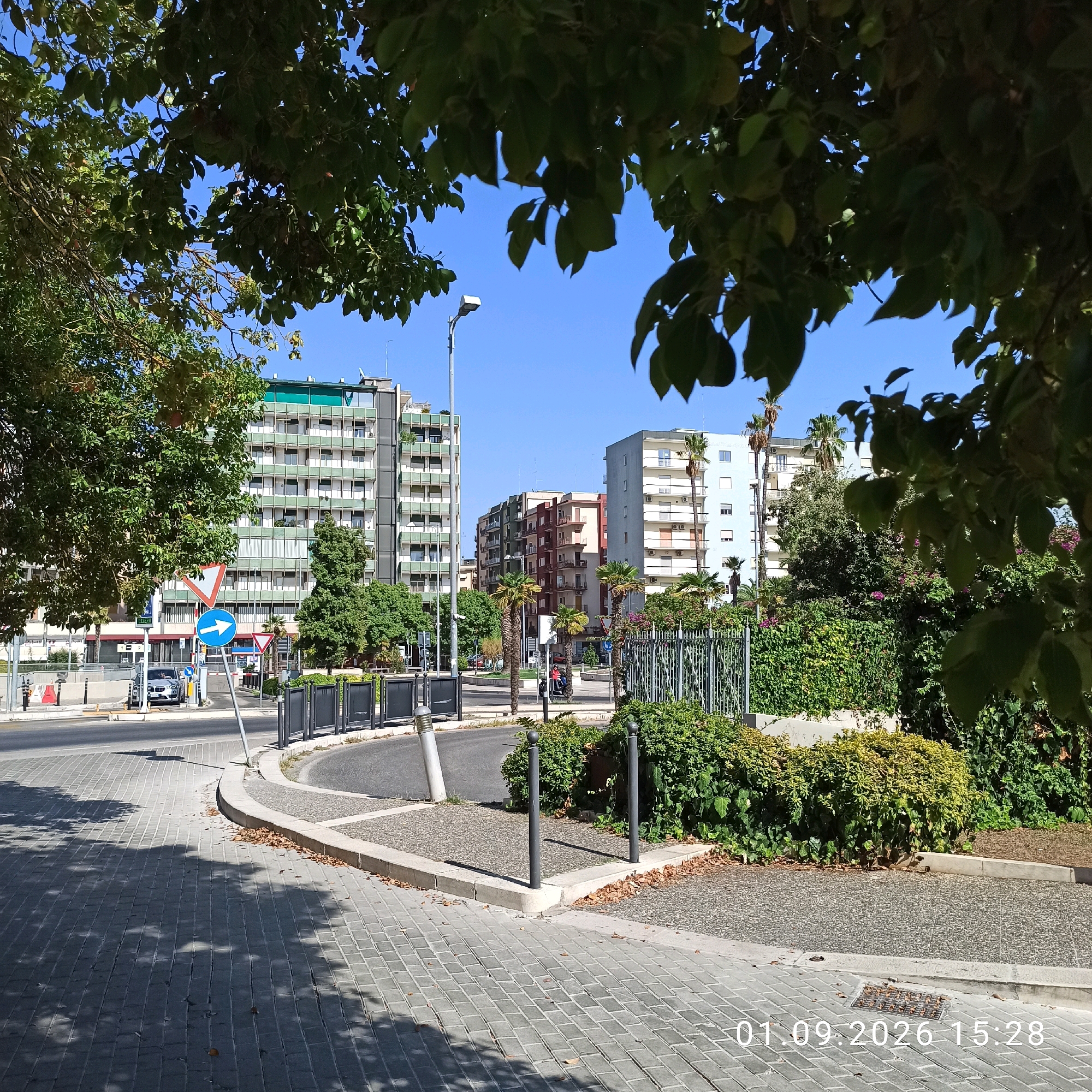 The image depicts a street scene in Bari, Italy, on a sunny day. The foreground is a paved pedestrian walkway with shadows from overhead trees. A curved road with a single lane is visible, bordered by a low concrete curb and a gravelly area with landscaping. Behind the landscaping is a metal fence.
In the mid-ground, traffic signs are present. One sign is a blue circular sign with a white arrow pointing to the right, indicating a mandatory direction. Another is an inverted red triangle with a white center, a yield sign. A silver SUV is parked near the intersection. Buildings line the background, appearing to be apartment complexes or office buildings. They vary in height and architectural style, with several featuring balconies and green trim. Palm trees and other greenery are scattered throughout the scene.
The sky is clear and blue. A street lamp is visible above the road. The date "01.09.2026 15:28" is superimposed on the lower right corner of the image.