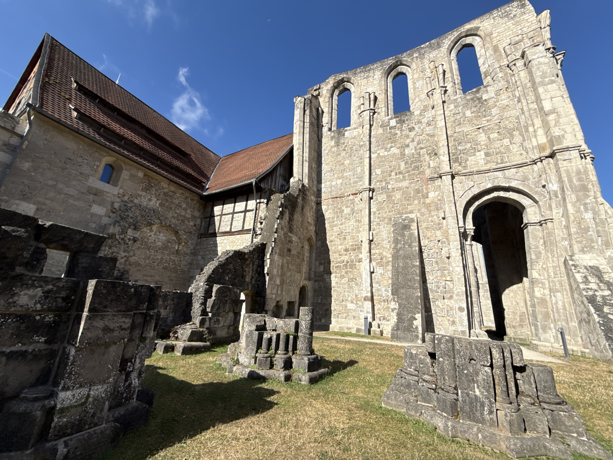 This is a wide-angle, eye-level outdoor photograph of the ruins of Walkenried Abbey in Walkenried, Germany. The sky is a clear, bright blue with a few wispy white clouds. The ruins, made of light-colored stone, dominate the right side of the frame. A large section of a wall stands tall with multiple arched window openings at the top, revealing the sky beyond. Below these openings, there are remnants of an arched doorway. To the left of the tall wall, a section of a more intact building with a red-tiled roof and stone walls is visible. This building has a smaller arched window. In the foreground and mid-ground, there are several scattered piles of weathered stone blocks, some resembling architectural fragments or bases of columns. The ground is covered in dry, yellow-green grass, with shadows cast by the stone structures. The overall impression is one of historical grandeur and decay under a clear sky.