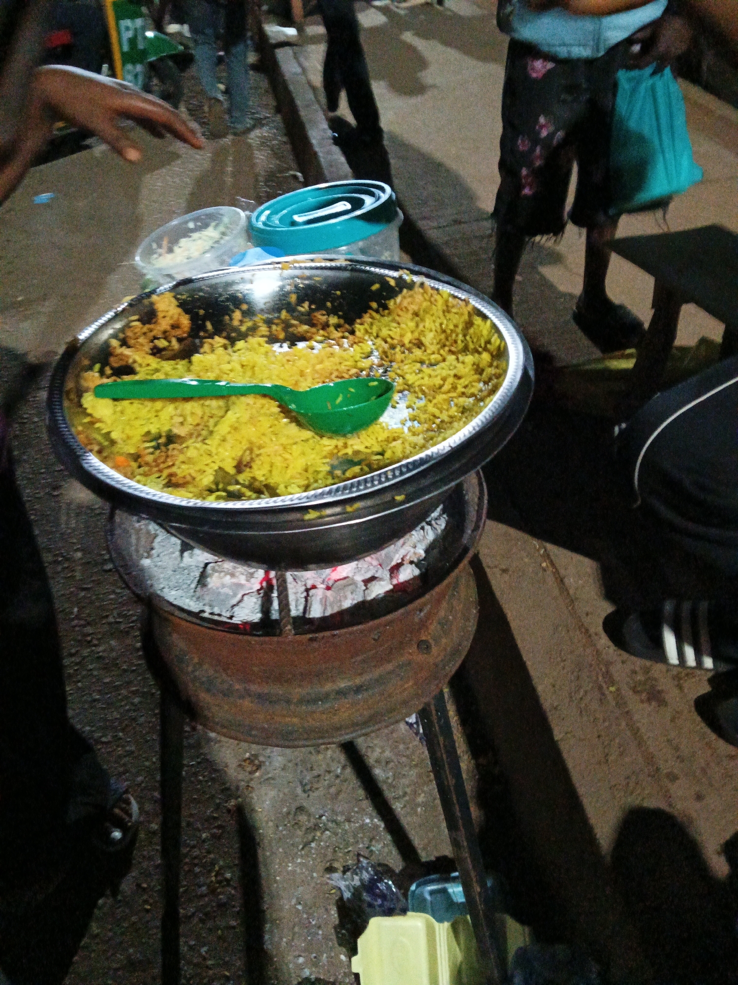 In Bukuru, Nigeria, a scene unfolds at ground level, illuminated by ambient light. A large, round metal tray, filled with a generous portion of yellow rice, is positioned atop a rusty, cylindrical metal brazier containing glowing red coals. A vibrant green plastic spoon rests on top of the rice. To the left of the tray, a clear plastic container holds a small amount of shredded vegetables, and next to it sits a teal-colored collapsible container.
In the background, slightly out of focus, are several individuals. One person, wearing patterned shorts and a blue top, holds a turquoise bag. Another person's hand, dark-skinned, is visible in the upper left corner, reaching down. The ground is paved with rough, dark material. The overall atmosphere suggests an outdoor food stall or market setting.