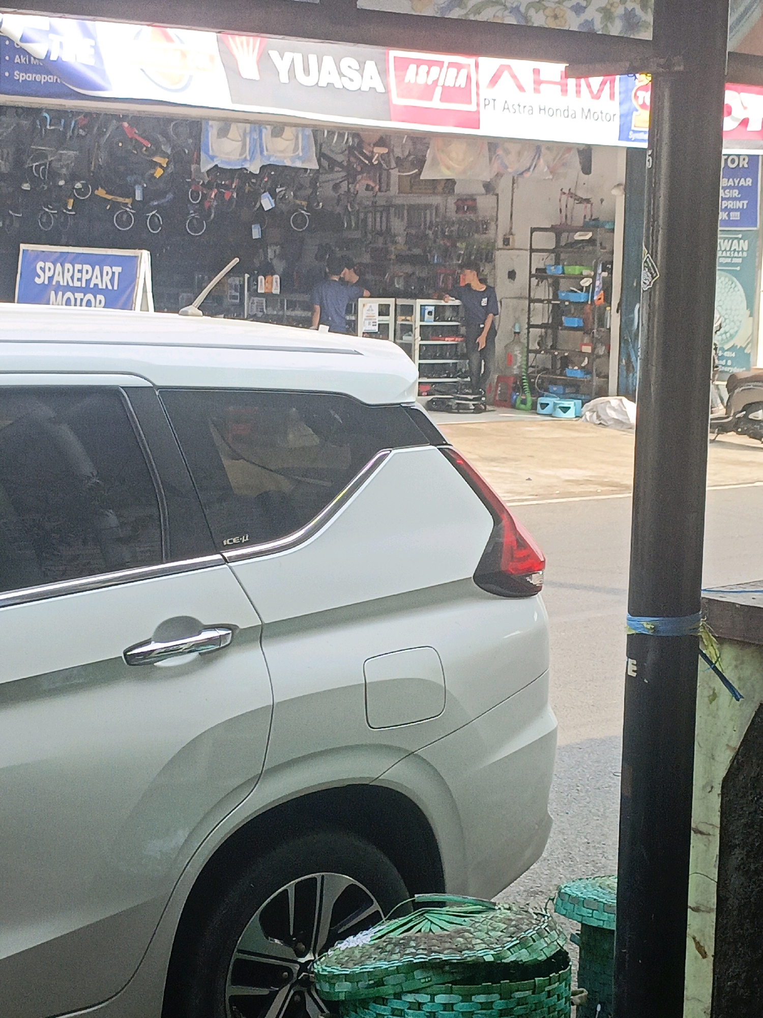 The image shows a white car parked in front of a store in Probolinggo, Indonesia. The store appears to be a spare parts shop, as indicated by the "SPAREPART MOTOR" sign. There are several people inside the store, which is stocked with various automotive parts. The street in front of the store is paved, and there are some plants and a lamppost visible. The car is a modern SUV, and it is parked parallel to the curb. The overall scene is a typical street view in a commercial area of a city in Indonesia.