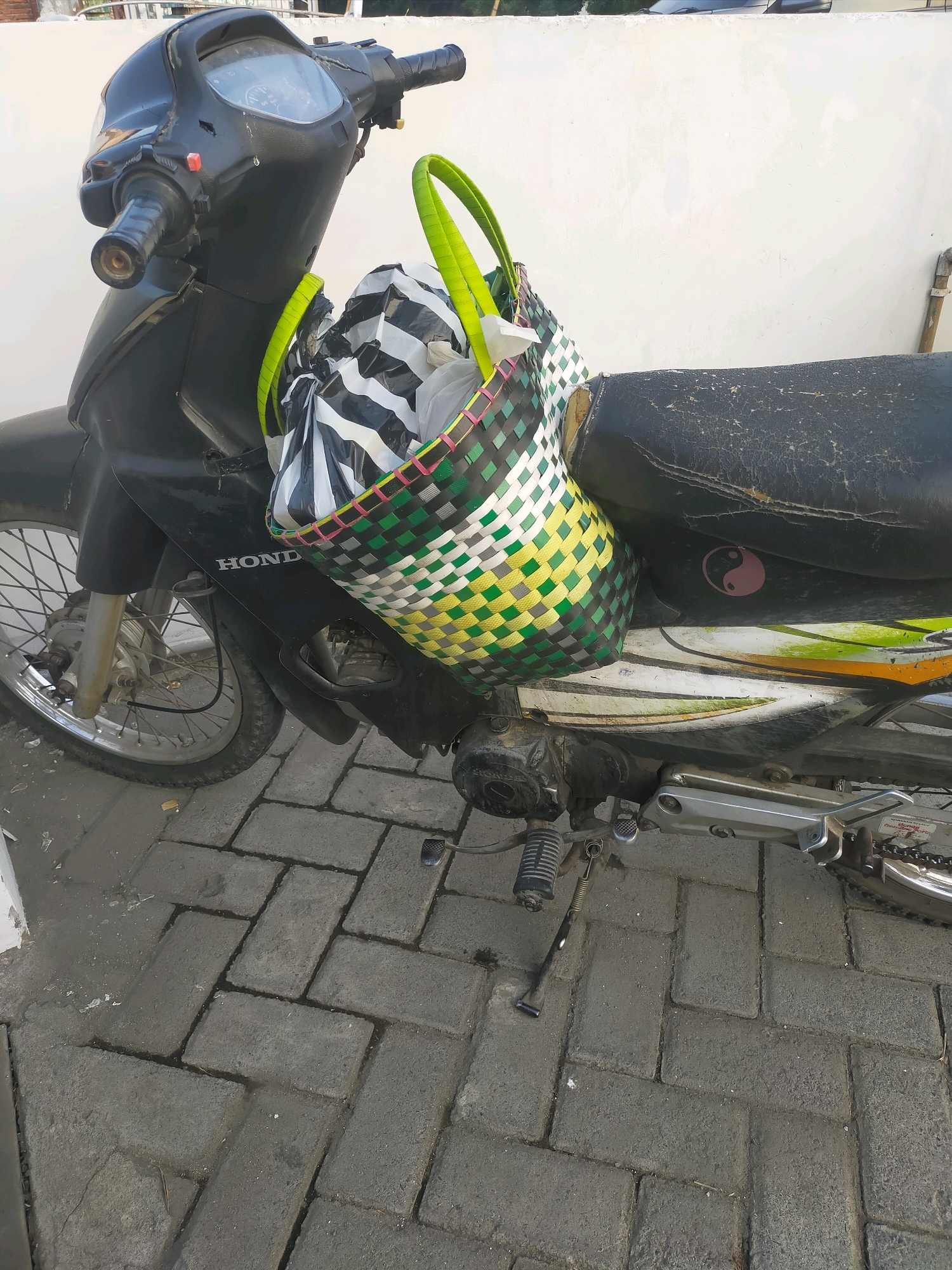 A black Honda scooter is parked on a brick-paved surface outdoors. A woven basket filled with items, including what appears to be a black and white striped plastic bag, is attached to the back of the scooter's seat with green straps. The scooter has a black seat with a pink yin-yang sticker. The scooter's front wheel and handlebars are visible. In the background, a white wall is present. The scene is located in the city of Puskesmas Keboan, Indonesia.