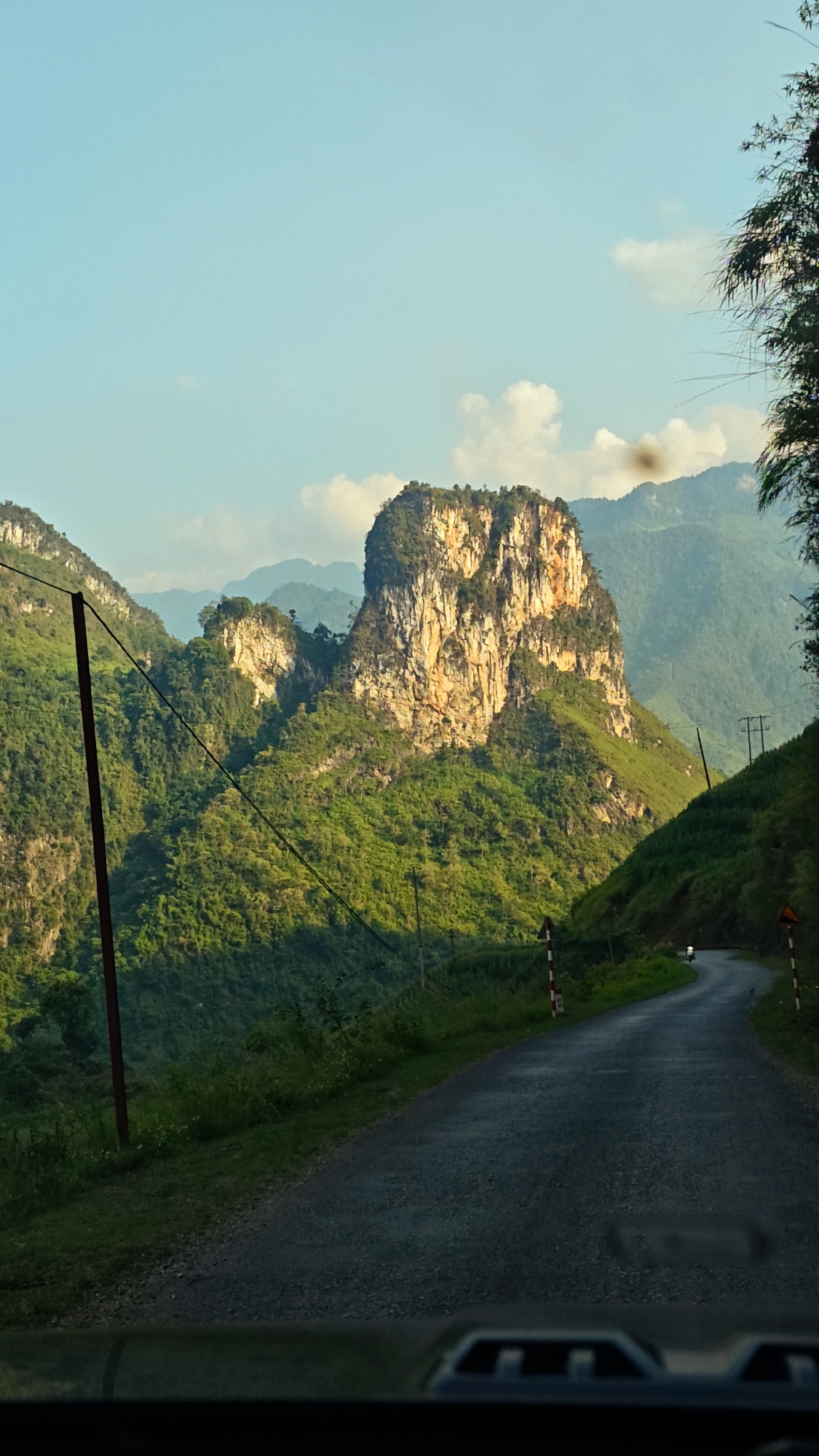 The image depicts a mountainous landscape in Đường Thượng Commune, Vietnam. A paved road curves through the terrain, with lush green vegetation covering the hillsides. In the background, a prominent rock formation dominates the scene, partially illuminated by sunlight. Power lines run along the road. A motorcycle is visible on the road in the distance. The perspective suggests the photo was taken from inside a vehicle.