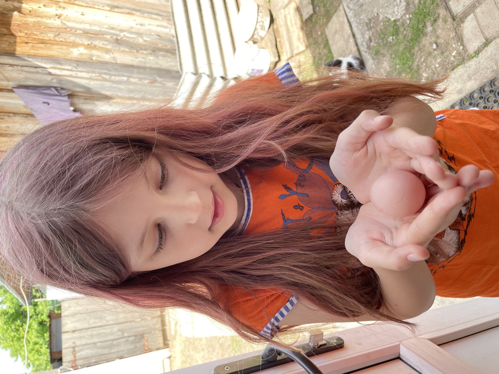 A young girl with dyed reddish-pink hair, wearing an orange t-shirt with a blue striped collar, is holding a pale pink egg in her hands. She is looking down at the egg with a slight smile. The background includes a wooden fence, foliage, and a partially visible black and white animal. The girl appears to be standing indoors, with a white frame structure visible at the bottom of the image. The location of the scene is unknown, as it's not possible to identify the city of Elokh, Russia, from the information provided in the image.
