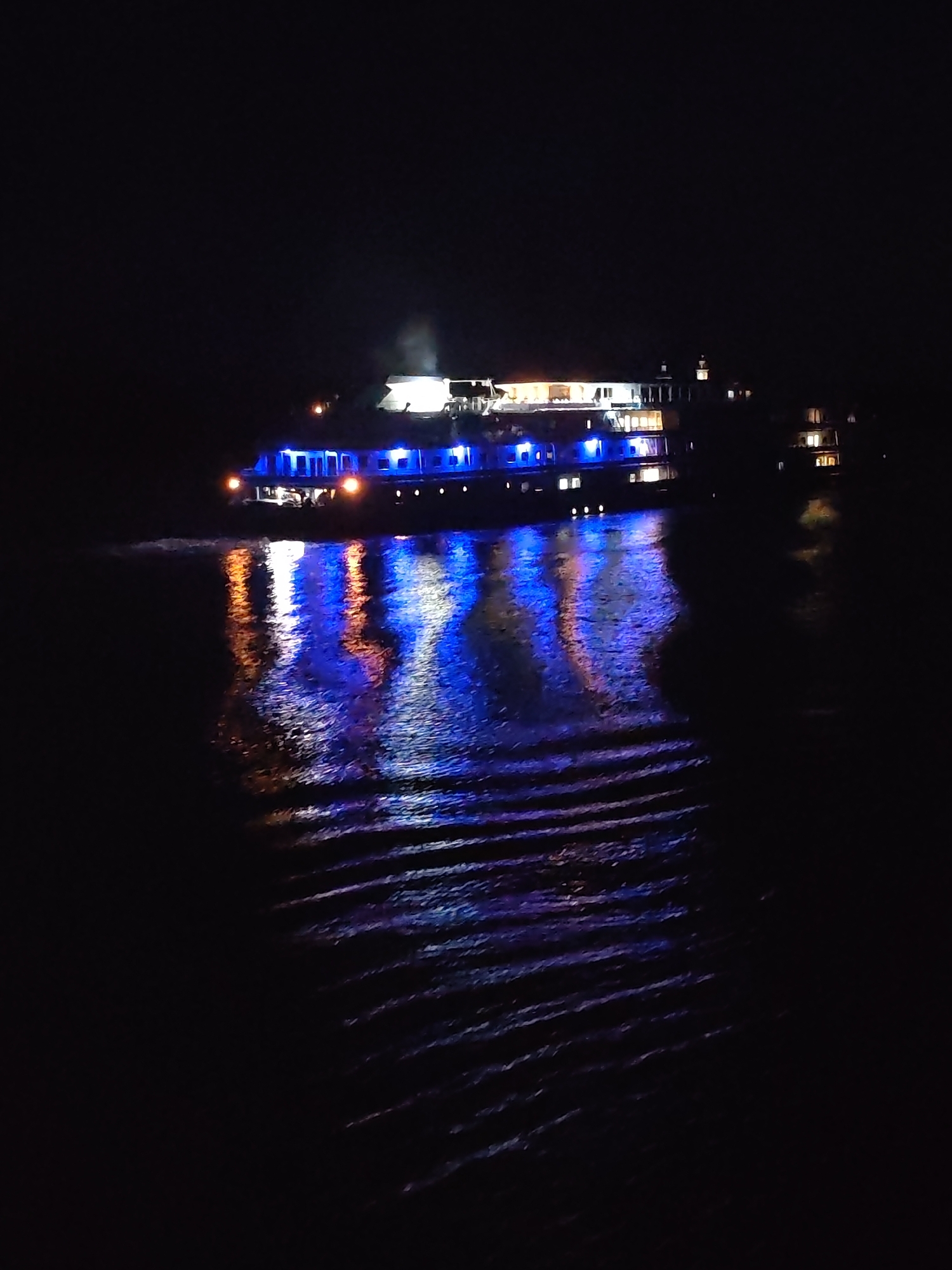 The image depicts a riverboat at night, likely on a body of water near Tikhonovo, Russia. The boat is lit with blue and white lights, which reflect in the water below. The wake of the boat is visible in the water, and there are lights from buildings on the shoreline in the distance. The overall scene is dark, with the boat and its reflections providing the primary sources of light.