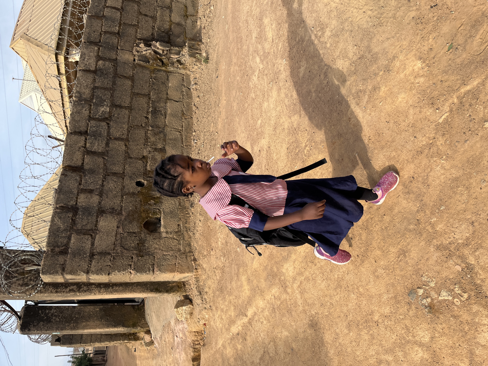 A young African girl in a school uniform stands outdoors on a reddish-brown dirt ground. She has a lollipop in her mouth and is wearing a black backpack and pink sneakers. The environment features a brick building with barbed wire on top. Shadows are cast on the ground, suggesting strong sunlight. The location is speculated to be in Jos, Nigeria.