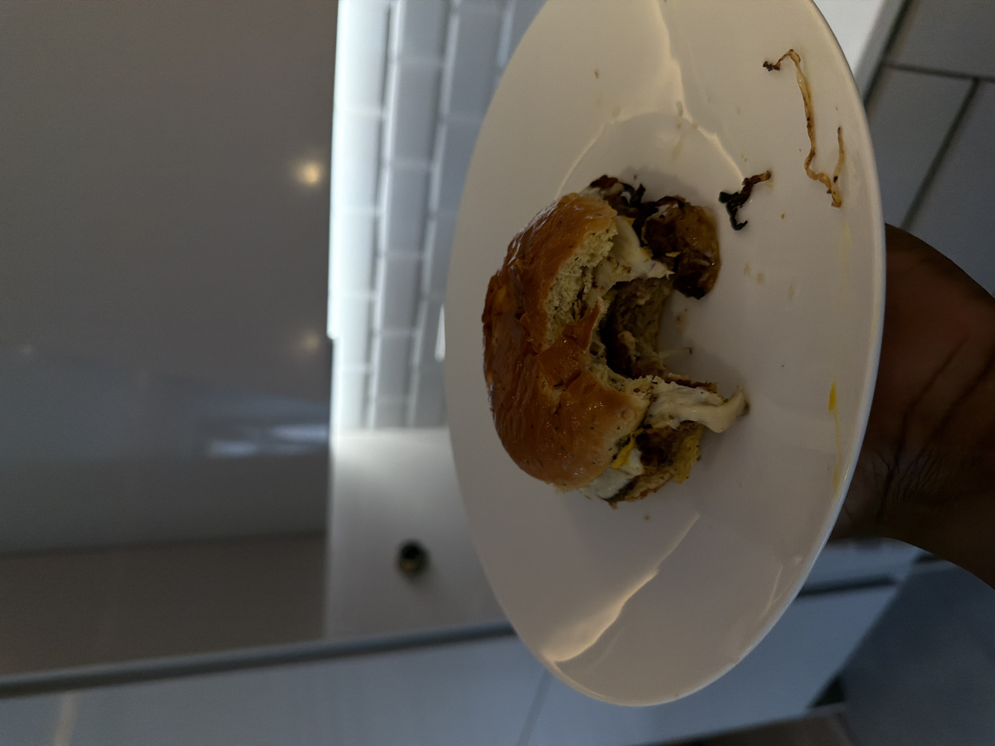 The image displays a close-up, tilted shot of a human hand holding a white plate with a partially eaten burger. The burger features a golden-brown bun, a visible meat patty, melted white cheese or sauce, and dark, stringy components, likely caramelized onions or fried shallots. Food residue, including yellowish stains and small dark fragments, is scattered on the white plate.
The hand holding the plate appears to have a dark skin tone. The scene is set indoors, with a bright, minimalist background. A large, light-colored vertical surface, possibly a wall with rectangular panels or tiles, is prominent behind the plate. Below this, there's a darker, reflective surface that could be a counter or floor. In the lower-left background, a small, dark circular object resembling a camera lens or sensor is visible. The artificial lighting suggests the image was taken inside, possibly in a modern kitchen, cafe, or dining area in London, United Kingdom. There is no visible text in the image.