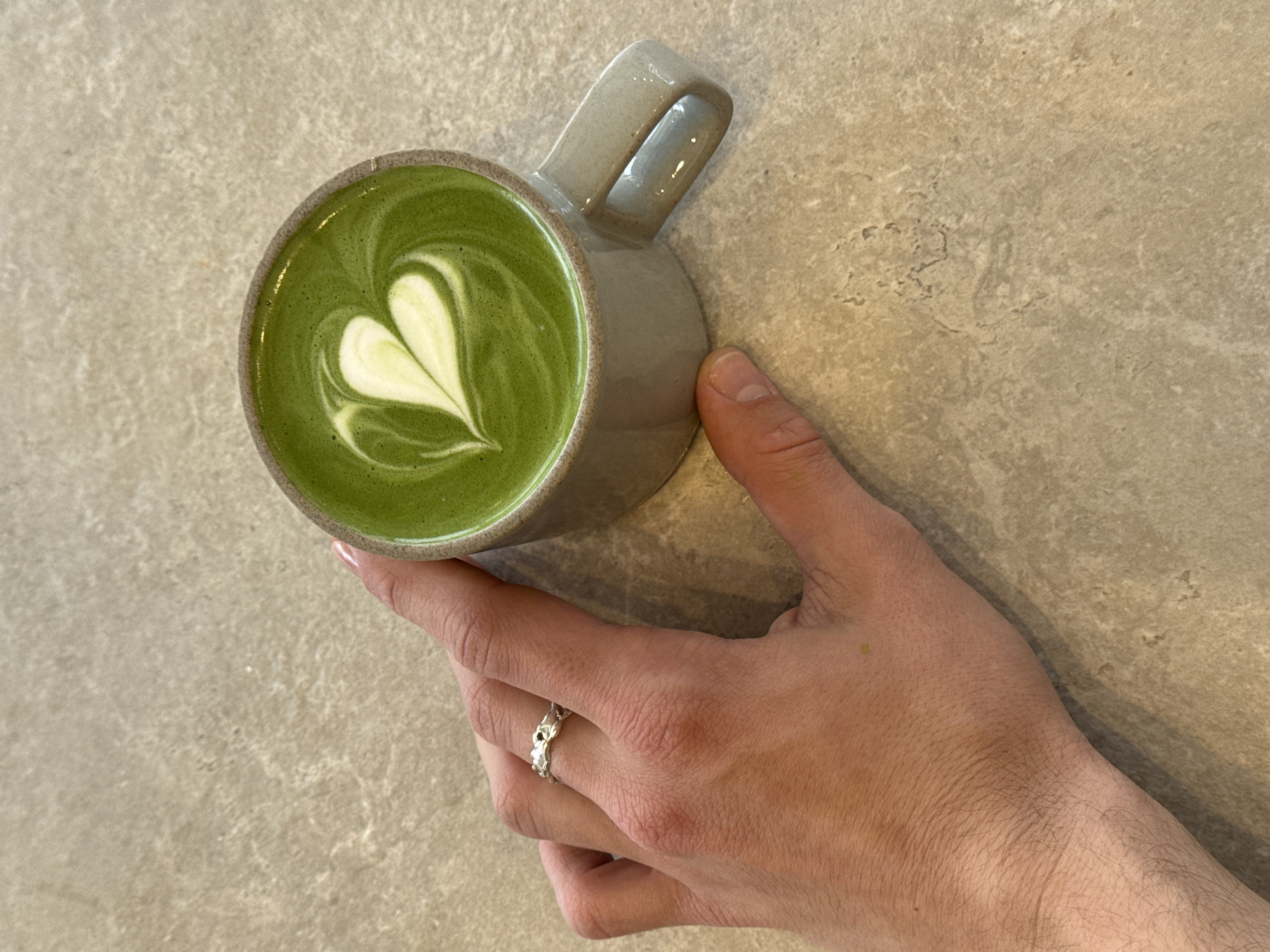 A hand holds a mug of matcha latte, with a heart-shaped foam design, on a beige, slightly textured surface. The hand is wearing a silver ring on the fourth finger. No information on location or environment is discernable.
