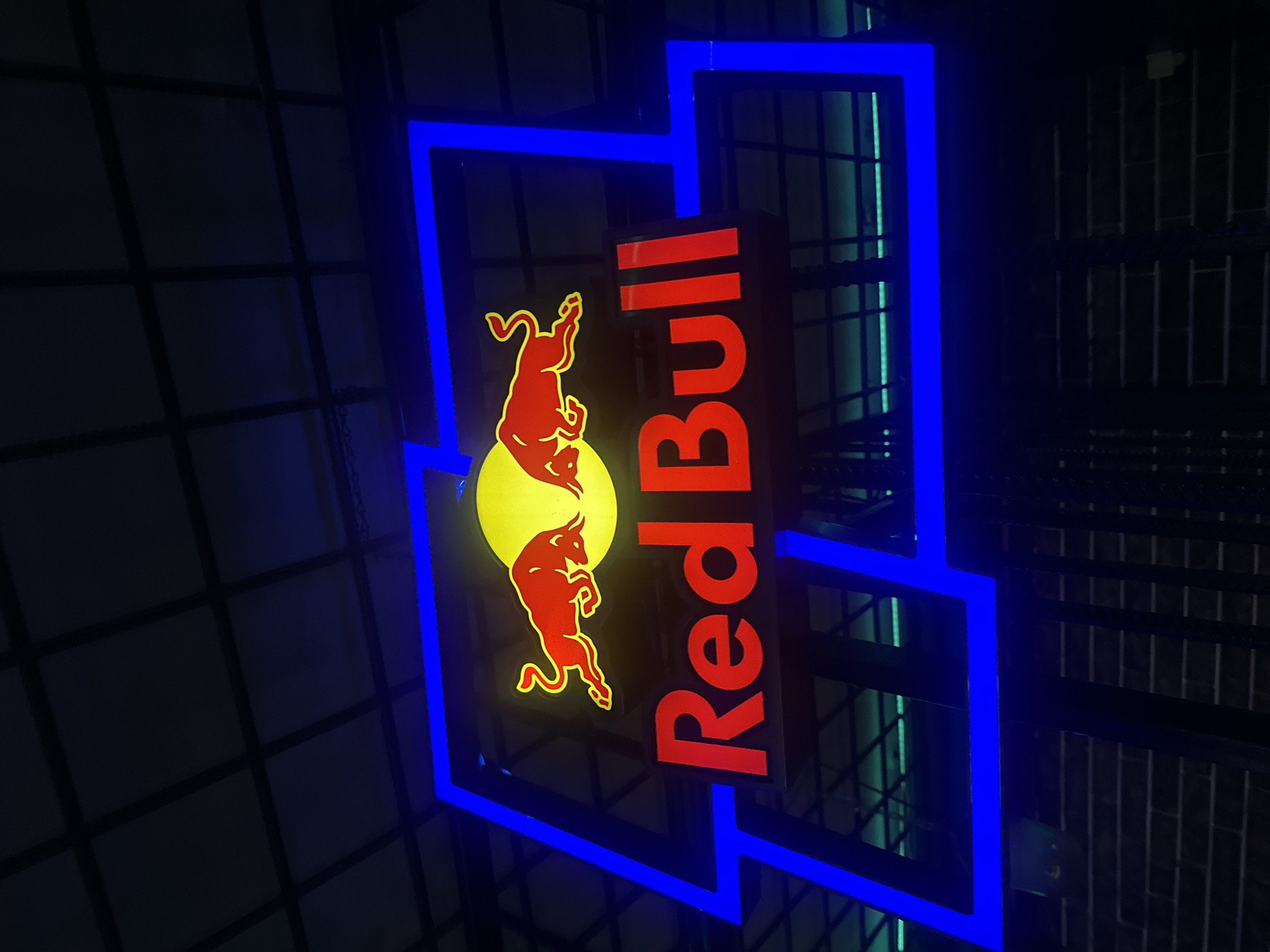 A nighttime shot of a "Red Bull" sign with two red bulls facing each other against a yellow circular background. The sign is framed by a blue, geometric outline. The background suggests an indoor environment with a grid-like structure. Yerevan, Armenia is not evident in the image.