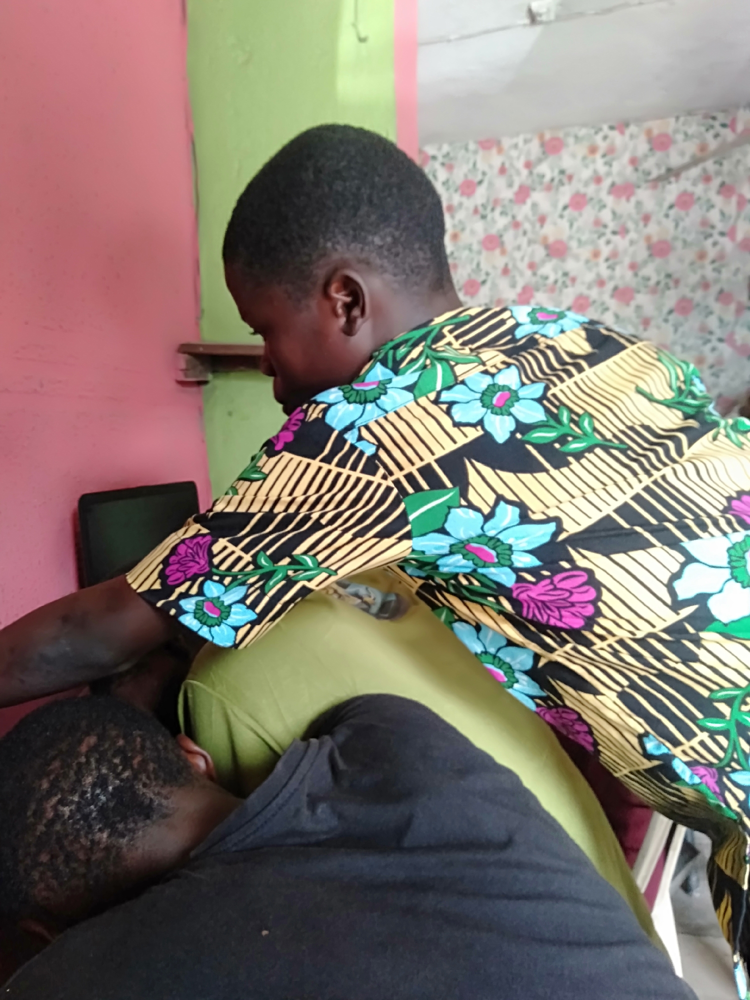 Two individuals are captured indoors, likely in a home setting in Shangisha, Nigeria. The primary subject, positioned in the center, is seen from the back and side, wearing a vibrant short-sleeved shirt adorned with a black, yellow, blue, green, and pink floral and striped pattern. This person has short, dark hair and is leaning forward, with their arm extended towards a dark-colored laptop or tablet visible on a surface to the left.
Partially visible in the lower left, from the back of their head and shoulder, is a second individual wearing a black top. This person also has short, dark, curly hair and appears to be resting or leaning against a light green cushion or pillow positioned between the two. The activity suggests a shared engagement, possibly focused on the electronic device.
The background features contrasting wall colors: a vibrant pink on the left and a lighter green on the right. A floral wallpaper, predominantly white with pink rose patterns, is visible on the far right wall. A small wooden shelf is mounted on the wall behind the main subject. The lighting indicates it is daytime. No text is visible in the image. The scene conveys a calm, collaborative, or focused interaction between the two individuals.