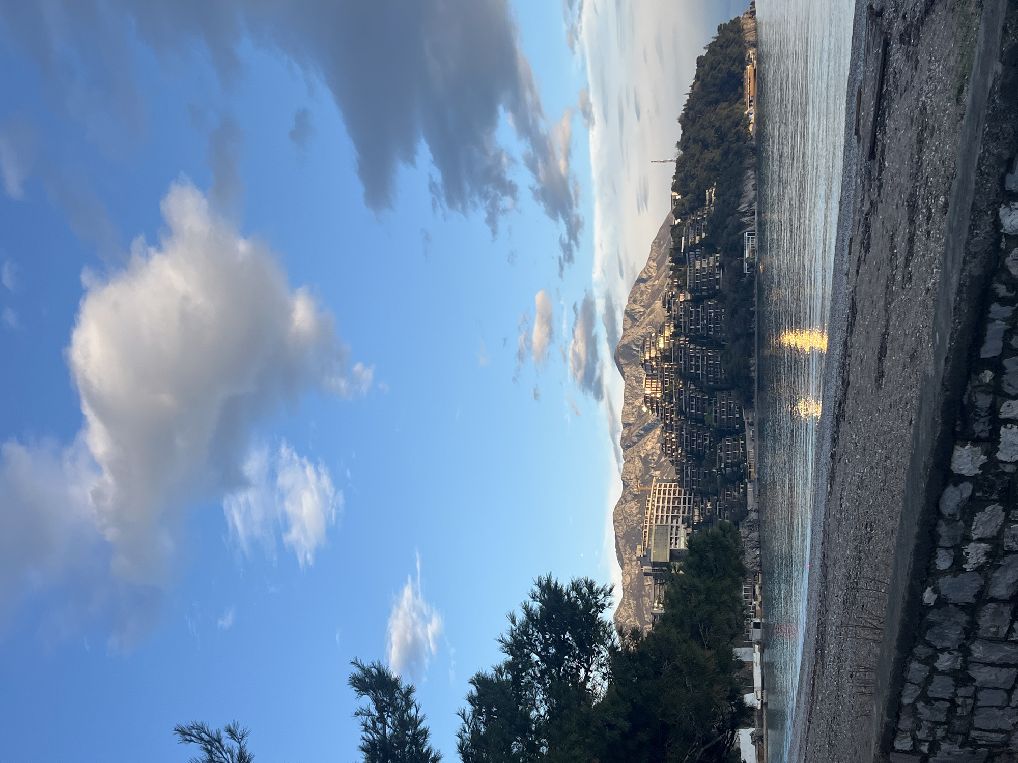 The image depicts a coastal scene in Budva, Montenegro, with a mix of natural and built elements. The sky is predominantly blue with scattered cumulus clouds. A mountain range rises in the background, its slopes covered in buildings. The sea is visible, reflecting sunlight. In the foreground, there are trees and a concrete walkway. The scene appears to be captured at dawn or dusk. No people are visible.