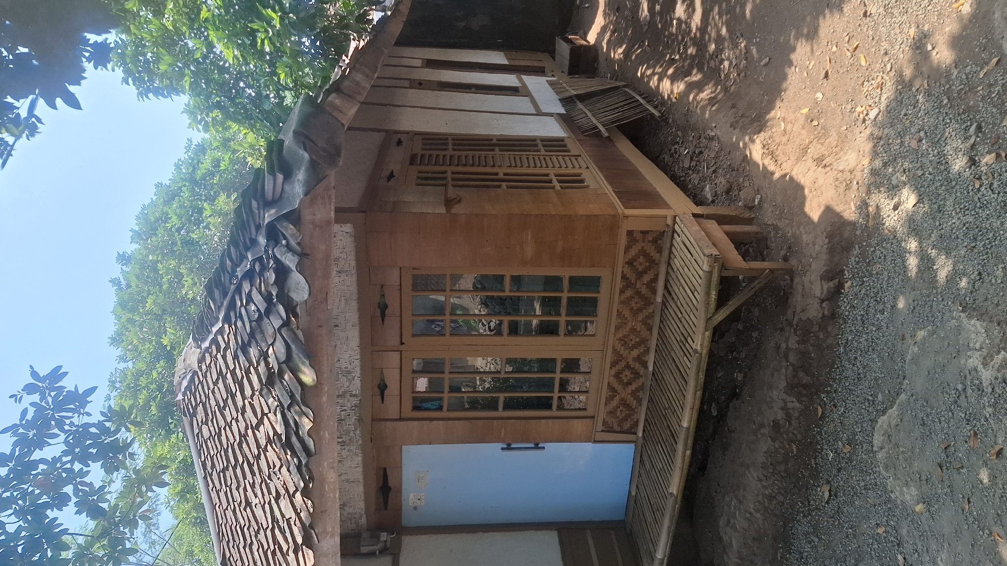 The image depicts a small, traditional wooden house situated outdoors in a rural or semi-rural setting, potentially in Cianjur, Indonesia, given the architectural style and vegetation. The house features a prominent tiled roof, which appears weathered. The walls are constructed from a combination of wood and what looks like woven bamboo or matting. A light blue door is visible on the left side of the house. Adjacent to the door and extending across the front of the house is a raised platform or veranda made of bamboo. Large, multi-paned windows with wooden frames are a significant feature of the house's facade. The area around the house is unpaved, with a mix of dirt, gravel, and scattered leaves. Lush green trees and foliage surround the structure, suggesting a natural environment. The sky is clear and bright blue, indicating daytime. The scene is static and presents a view of the exterior of the dwelling. There are no people visible in the image.