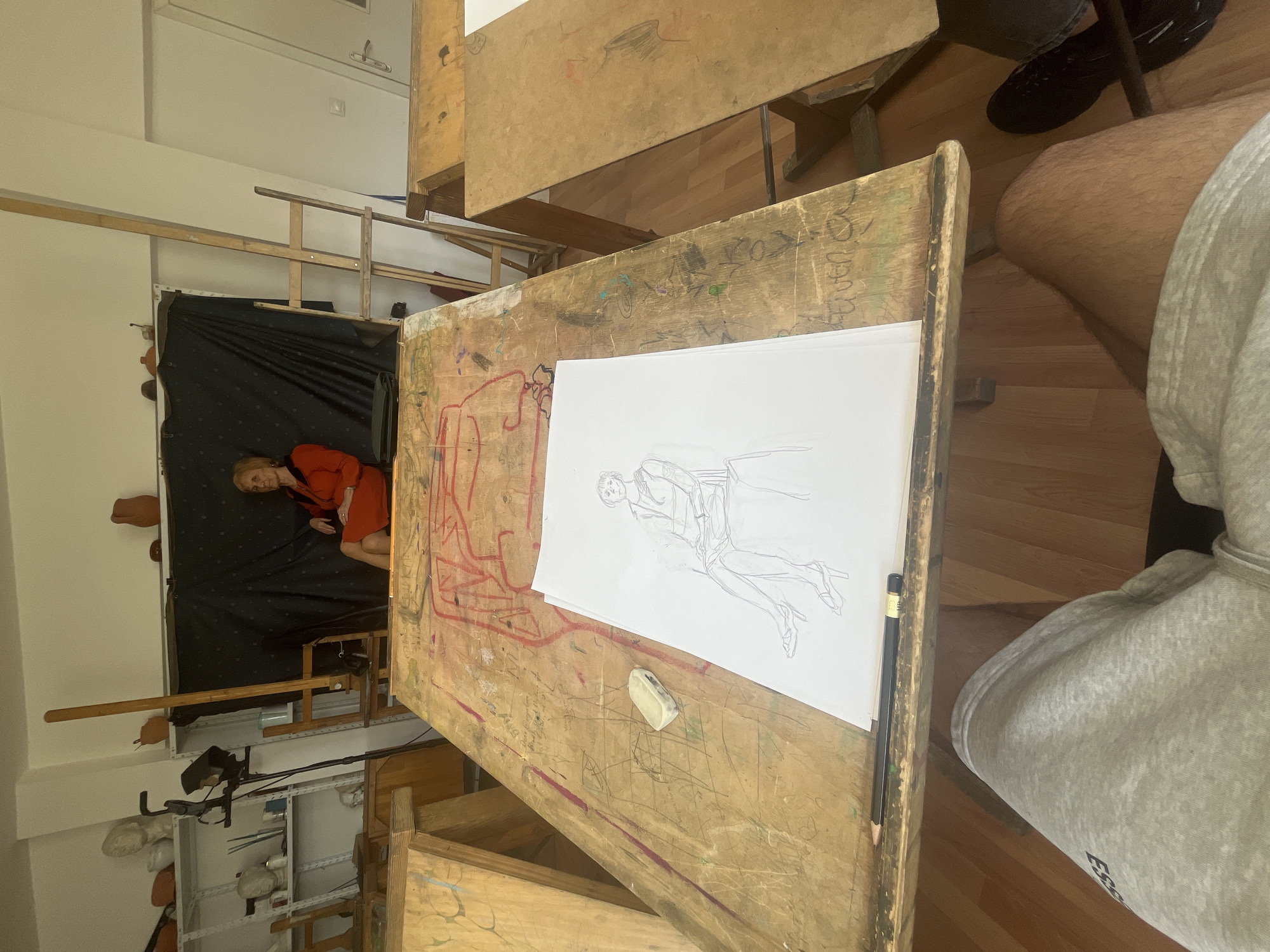 A medium-angle, slightly canted, interior shot shows an art class in progress. In the background, a woman in a red dress poses on a black backdrop. In the foreground, on a wooden drawing table, is a sketch of the model on white paper. Also on the table is a pencil and an eraser. A person wearing a gray sweatshirt sits at the table. The location appears to be a studio in Sofia, Bulgaria, given the subject matter and likely educational setting. There are other drawing tables visible. The room contains shelves with art supplies.