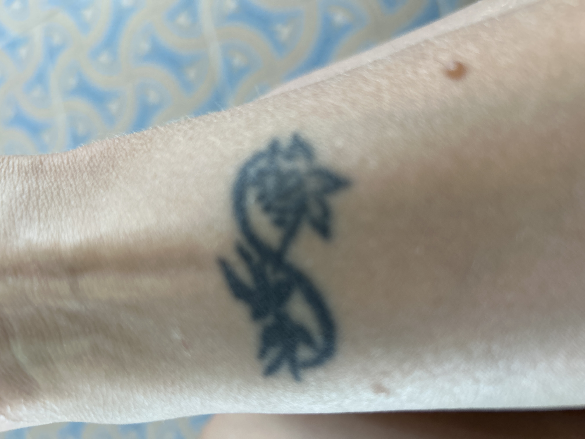 The image shows a tattoo on a person's forearm. The tattoo is black and appears to be an infinity symbol with floral elements. The background is a blurred patterned fabric. The image is taken in a close-up shot, focusing on the tattoo. There is no visible indication of the city of Gelendzhik, Russia, or any specific location within a city.