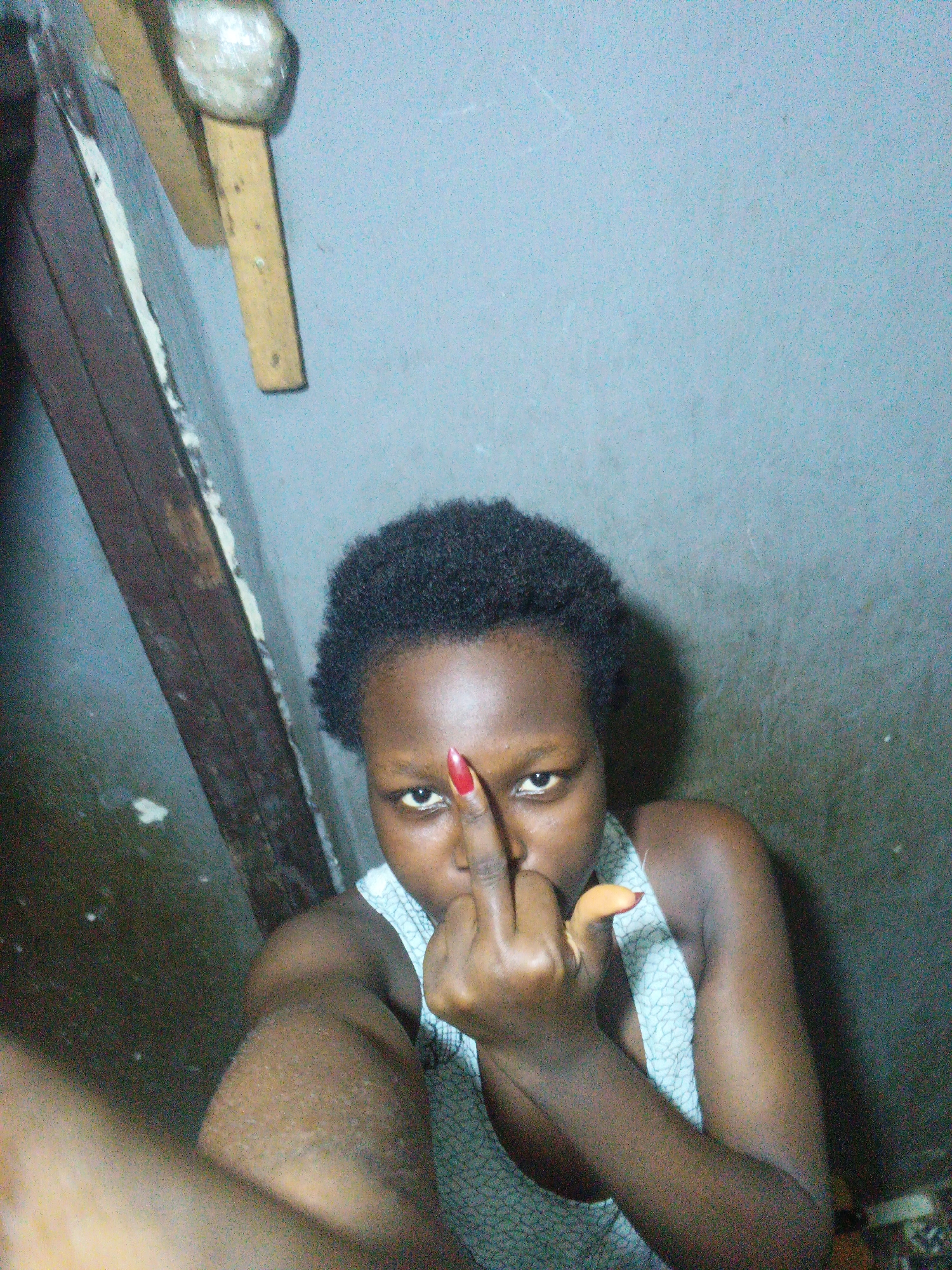 In Onitsha, Nigeria, a young woman with dark skin and short, black hair is captured in a selfie. She is wearing a grey and white patterned tank top. Her right hand is raised, and she is extending her middle finger towards the camera, with her fingernails painted red. The background appears to be an interior space with a grey, textured wall and a wooden structure to the left, which seems to be part of a doorway or frame. The lighting is dim, creating shadows and a grainy texture in the image. The floor area on the left shows signs of wear and potential dampness.