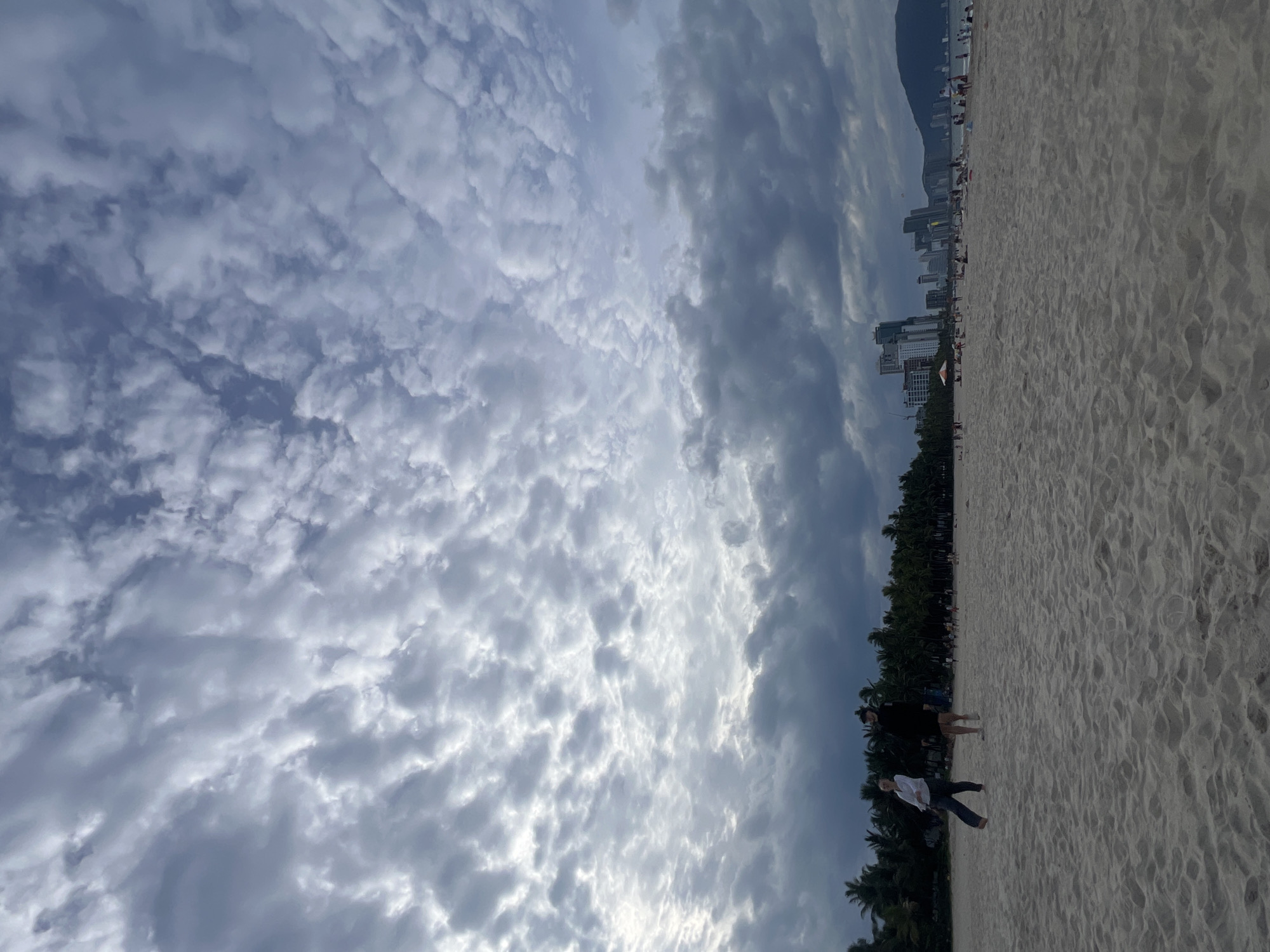 The image depicts a beach scene in Đà Nẵng, Vietnam, under a cloudy sky with a mix of dark and light formations. In the foreground, soft, light-colored sand stretches towards the horizon, showing footprints and texture. Two individuals are walking on the beach; one, on the left, wears a white top and dark pants, appearing to be mid-stride. The second person, slightly behind and to the right, is dressed in dark clothing and a cap.
Further back, a dense line of palm trees forms a dark green backdrop to the beach. Beyond the trees, a cityscape is visible, characterized by numerous modern high-rise buildings. Some of these buildings are tall and slender, reflecting the sky and surrounding light. A few smaller structures and an orange umbrella are also discernible on the beach among scattered people. The presence of buildings and the overall ambiance suggest a populated urban beach area within Đà Nẵng. The water is not clearly visible due to the angle and the distance. The overall lighting indicates it is likely late afternoon or early evening, given the dramatic cloud formations and subdued sunlight.