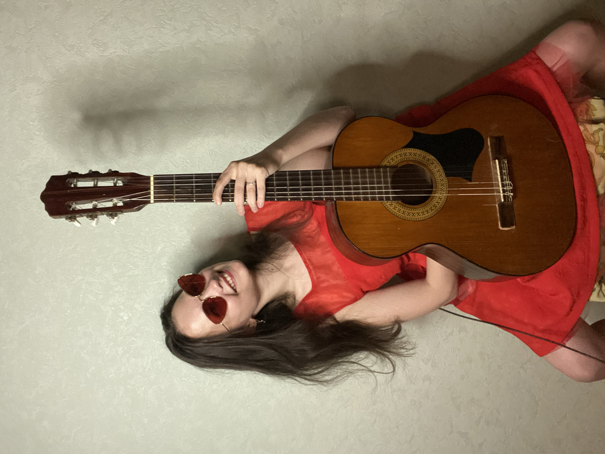 A young woman with long, dark hair and red, heart-shaped sunglasses is sitting and holding an acoustic guitar in front of a textured, light-colored wall. She is wearing a bright red dress with sheer sleeves. Her left arm is wrapped around the neck of the guitar, and her right hand is resting on the fretboard. She is smiling and looking upwards to her left. A thin black cable runs from behind the guitar, towards the lower left of the frame. The scene appears to be indoors, possibly in a residential setting. There is no identifiable information within the image to determine its location in the city of Chaikovsky, Russia.