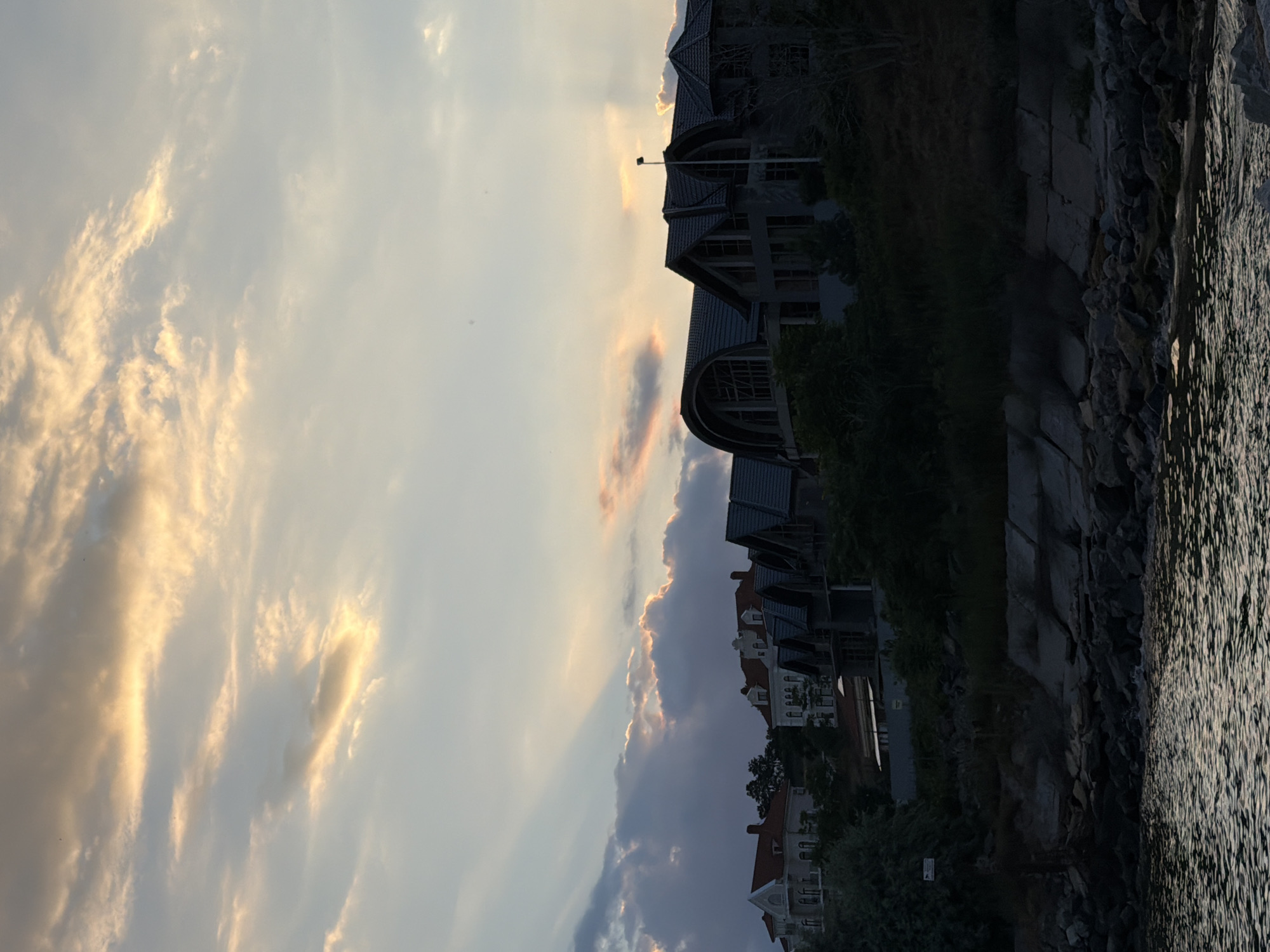 The image depicts a twilight scene featuring a row of houses situated on a hill overlooking a body of water. The sky is filled with dramatic clouds, illuminated by the setting sun, casting rays of light and warm hues across the sky. The houses vary in architectural style, with some appearing to be older with red tile roofs, and others more modern with dark, sloped roofs and large windows. The foreground is dominated by a rocky shoreline and calm water that reflects the sky's colors. The overall impression is one of a serene, picturesque evening in a coastal setting. The location is identified as Fontanka, Ukraine.