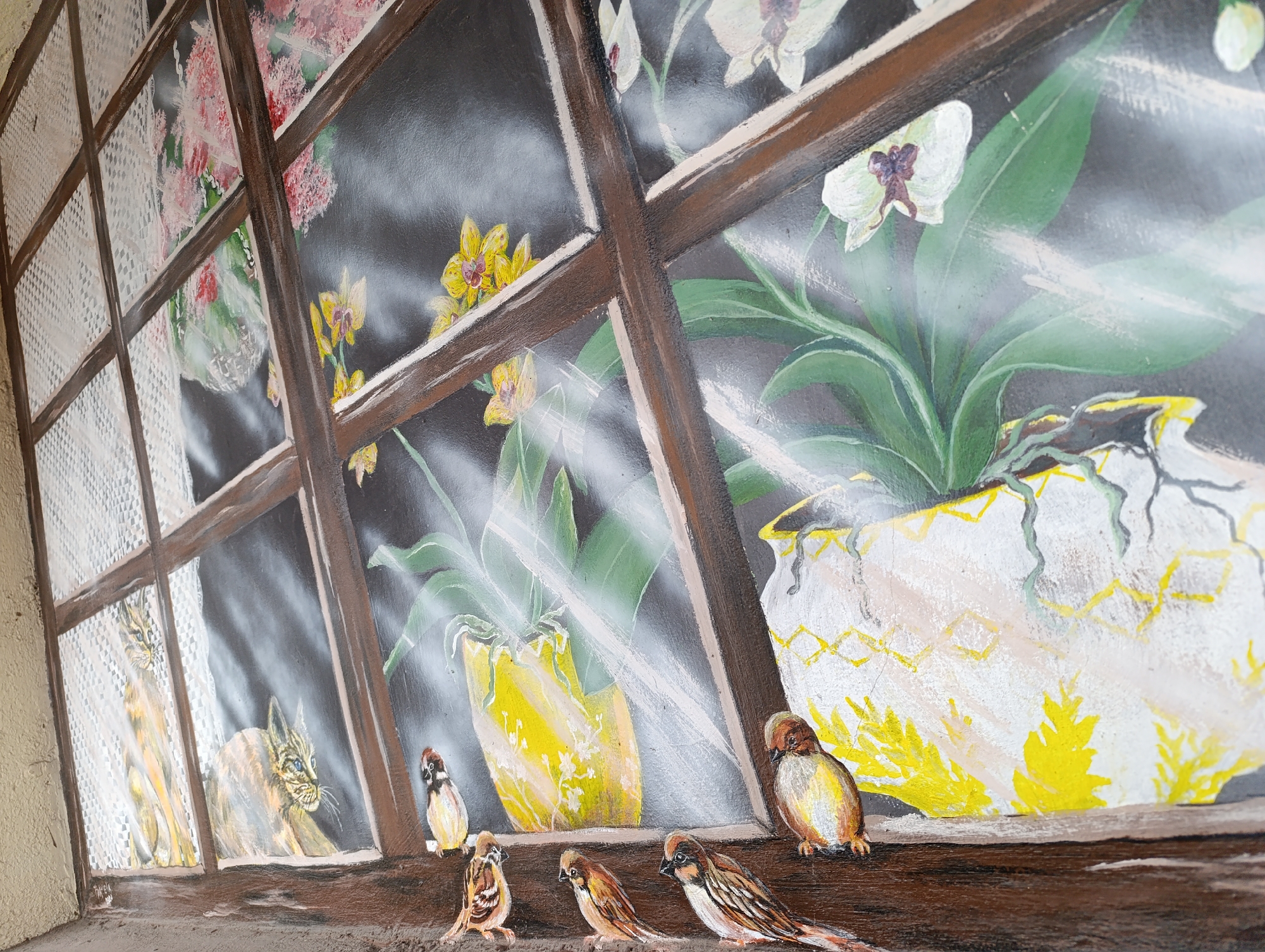 The image depicts a mural in Haskovo, Bulgaria, painted on an exterior wall. The mural simulates a window view, showing potted plants with yellow and white flowers on the windowsill. In front of the window on the sill are five brown sparrows. Through the window, two cats can be seen behind a lace curtain in the leftmost window pane. The window frame is painted brown, and the background behind the plants appears dark. The bottom of the wall is painted brown to simulate the sill.