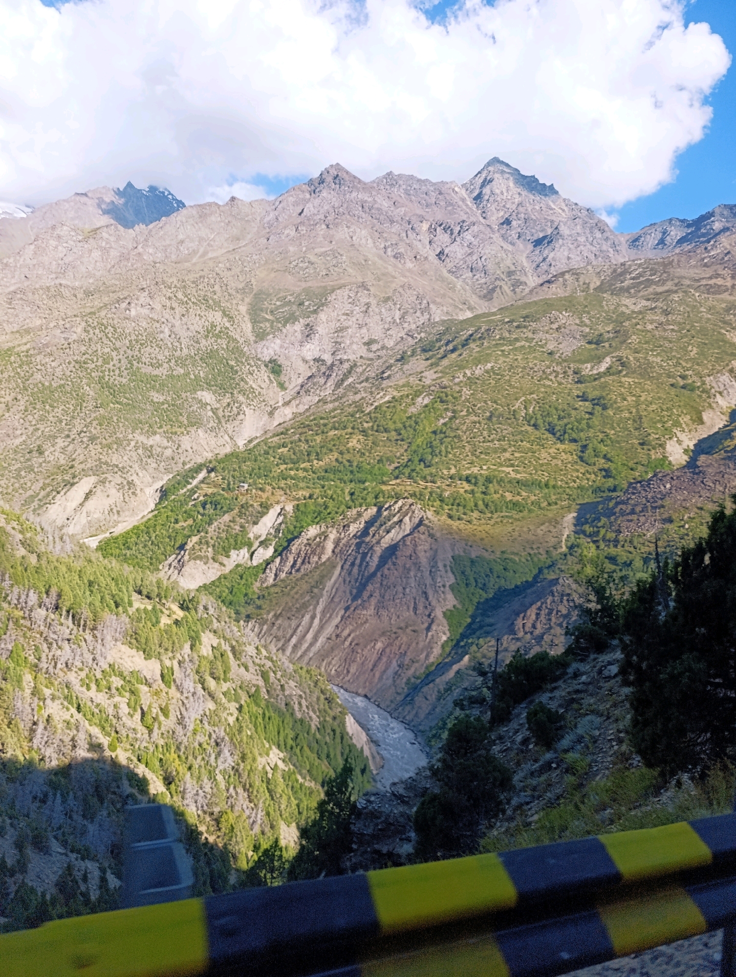 This is a landscape photograph captured from a high vantage point, overlooking a mountainous region. The scene is dominated by rugged, rocky peaks that rise majestically into a partly cloudy sky. The slopes of the mountains are covered with sparse vegetation, varying from dry, scrubby bushes to patches of green trees in the lower valleys. A river or stream flows through a deep gorge in the middle ground, its water appearing turbulent and grey. The light suggests it is daytime, with shadows cast across the landscape, indicating the sun's position. In the foreground, a portion of a safety barrier with yellow and black stripes is visible, suggesting the viewpoint is from a road or a viewing platform. The overall impression is one of a remote, natural environment with dramatic topography. The image does not contain any people or specific urban elements that would allow for pinpointing a location within the city of Stingri, India.
