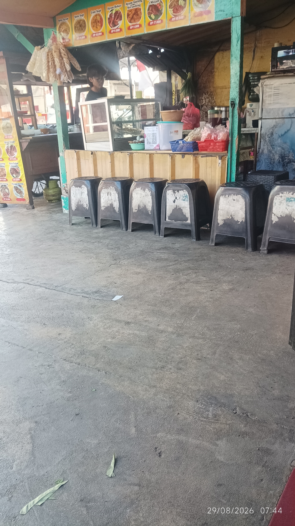 The image depicts an outdoor food stall in Tasikmalaya, Indonesia. The stall features a counter with food menu posters displayed above. Behind the counter, a person wearing glasses and a black shirt stands, possibly a vendor. Several plastic stools are neatly arranged in front of the counter. The ground is paved with concrete. To the left of the stall, a large green gas tank is visible, and a menu board is attached to a stand. Various food items and containers are present on the counter, including baskets filled with food products. The overall environment suggests a casual dining setting.