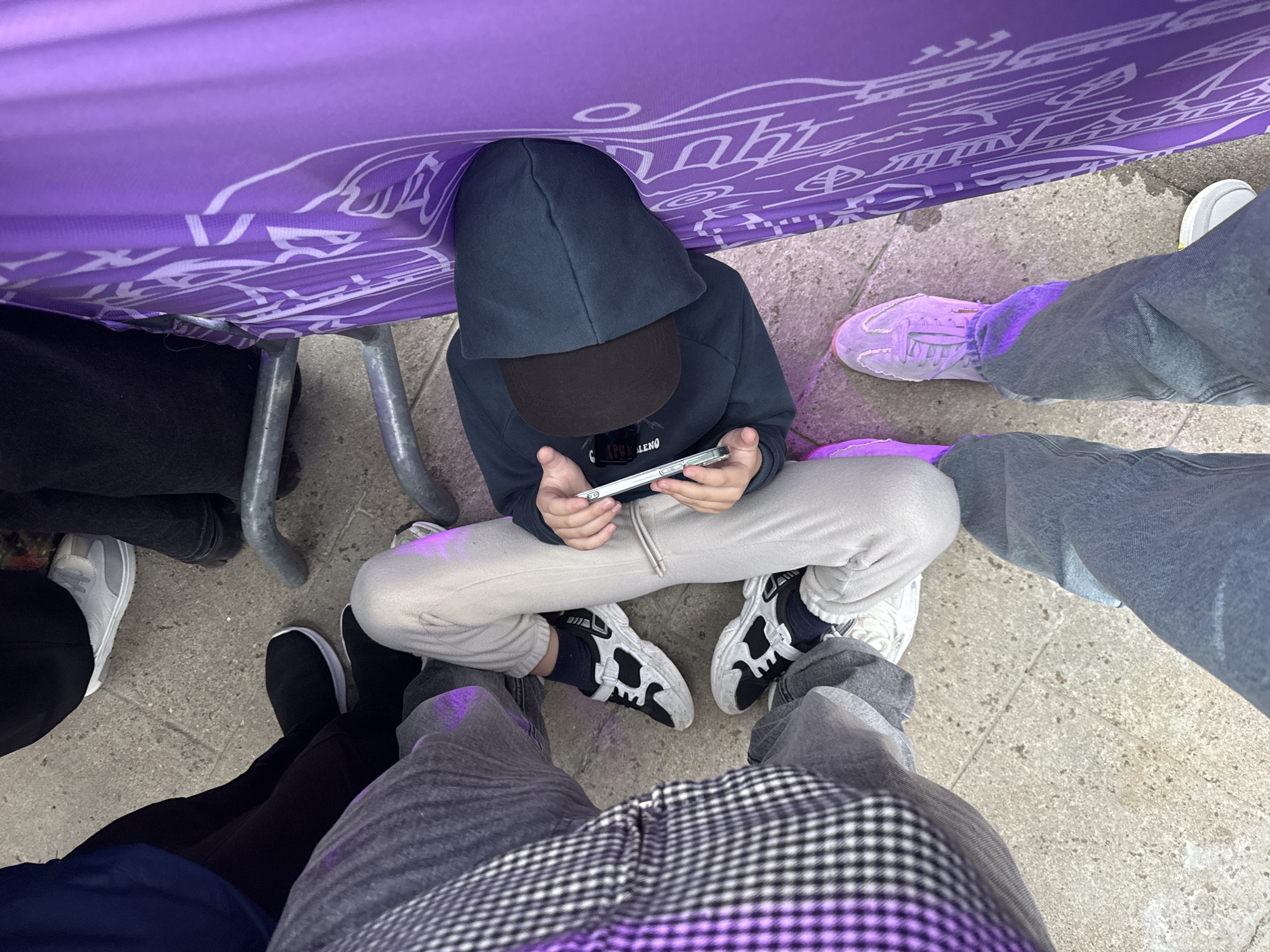 A high-angle shot shows a child in a dark blue hooded sweatshirt and black baseball cap sitting cross-legged on a concrete surface. The child is holding a white smartphone and appears to be looking at the screen. To the left of the child is a leg covered in black pants, with a gray sneaker visible. To the right are legs covered in gray pants, with lilac sneakers. A set of legs in gray-and-black checkered pants sit in the foreground of the frame. A purple surface printed with white graphics covers the top of the frame. The location is Sirius, Russia.