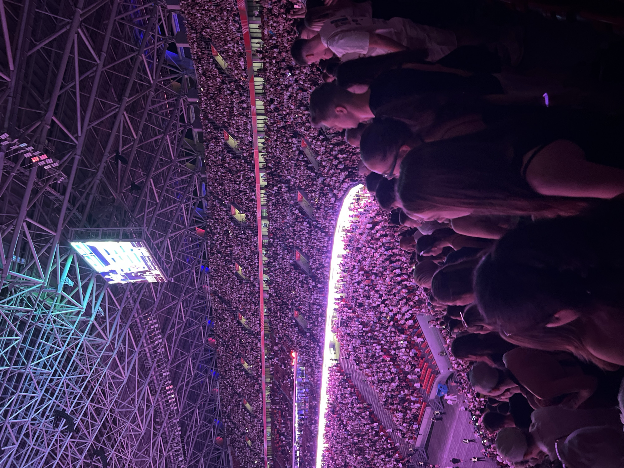 This image captures a wide view of a crowded stadium in Budapest, Hungary, bathed in vibrant purple and pink lighting. The stadium's complex metallic roof structure is visible overhead, with a large screen displaying content. Thousands of spectators fill the tiered seating, a sea of predominantly white shirts, creating a visually striking effect under the colored illumination. In the foreground, the heads and shoulders of several individuals are visible, suggesting the perspective of someone within the audience. The overall atmosphere appears to be that of a large-scale event, possibly a concert or sporting match, given the scale of the venue and the enthusiastic crowd. The scene is dynamic and immersive, with the lighting playing a significant role in defining the mood.