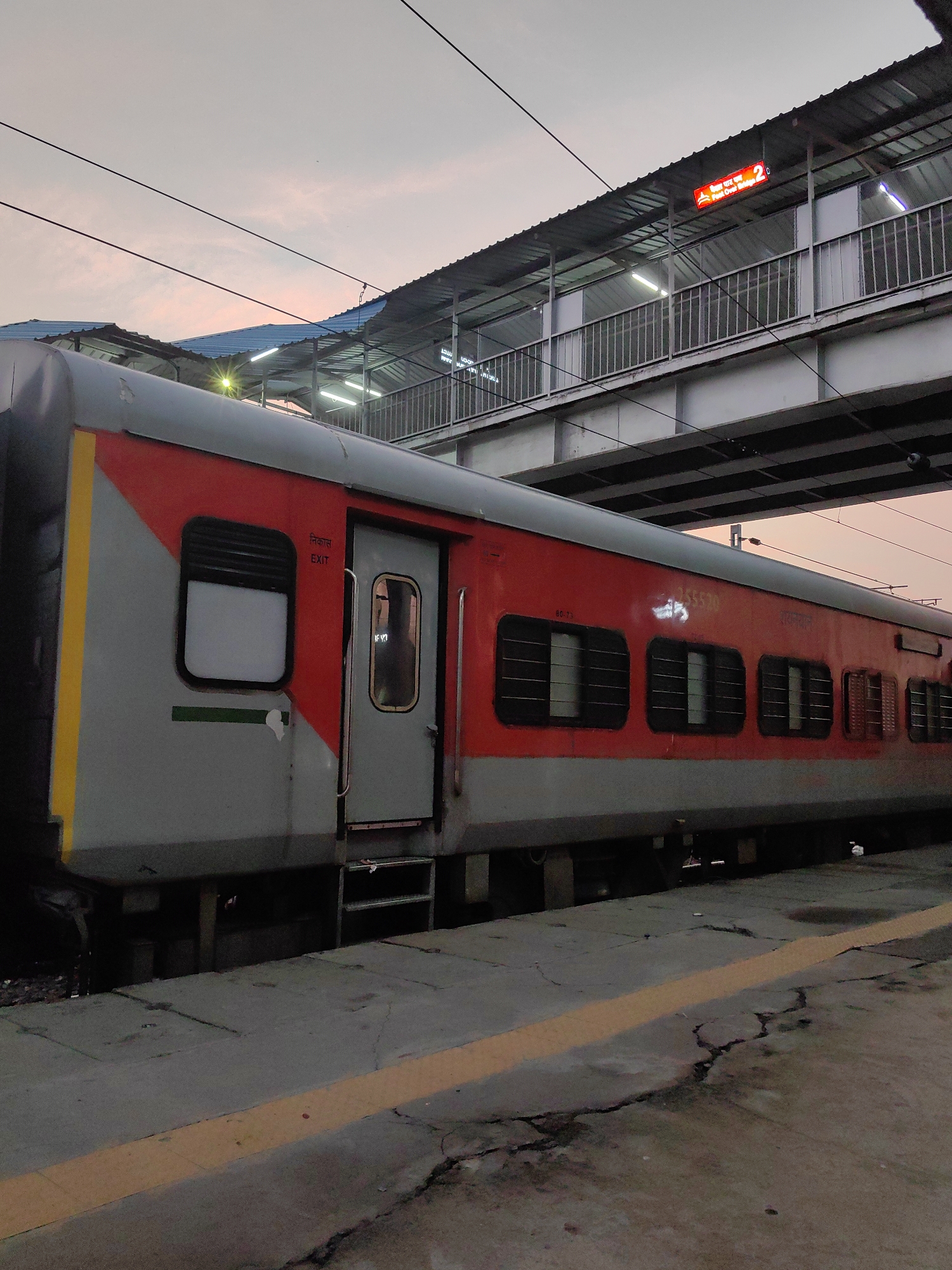 The image captures a train car at a railway station in Varanasi, India, under a twilight sky. The train is primarily red and gray, with yellow and green accents. A door labeled "EXIT" is visible on the left side of the train. Above the train, a pedestrian overpass with a metal roof and railings is present, illuminated by lights. A red sign with white text indicating "Platform 2" is attached to the overpass. The platform itself is made of concrete with a yellow warning line along its edge. The scene is static, with no people or activity evident. The environment suggests an urban railway setting.