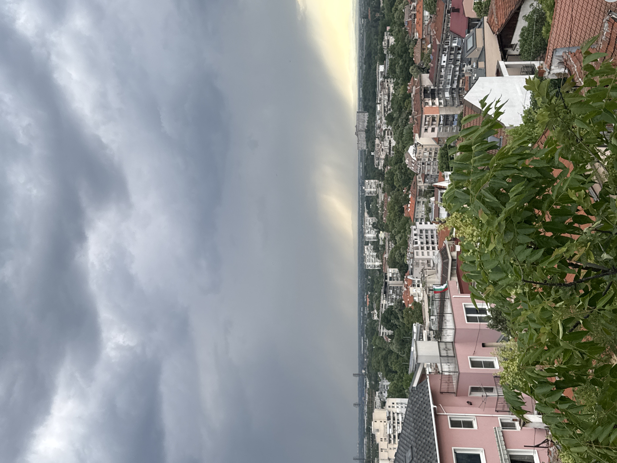 The image shows a view over Plovdiv, Bulgaria, under a stormy sky. The left side of the image is dominated by dark, grey storm clouds. A bright strip of light separates the storm from the city. The city consists of buildings, trees, and infrastructure. A pink building with a gray roof and white windows is visible in the lower right corner, partially obscured by a tree. Numerous other buildings of varying heights and colors are seen throughout the city.