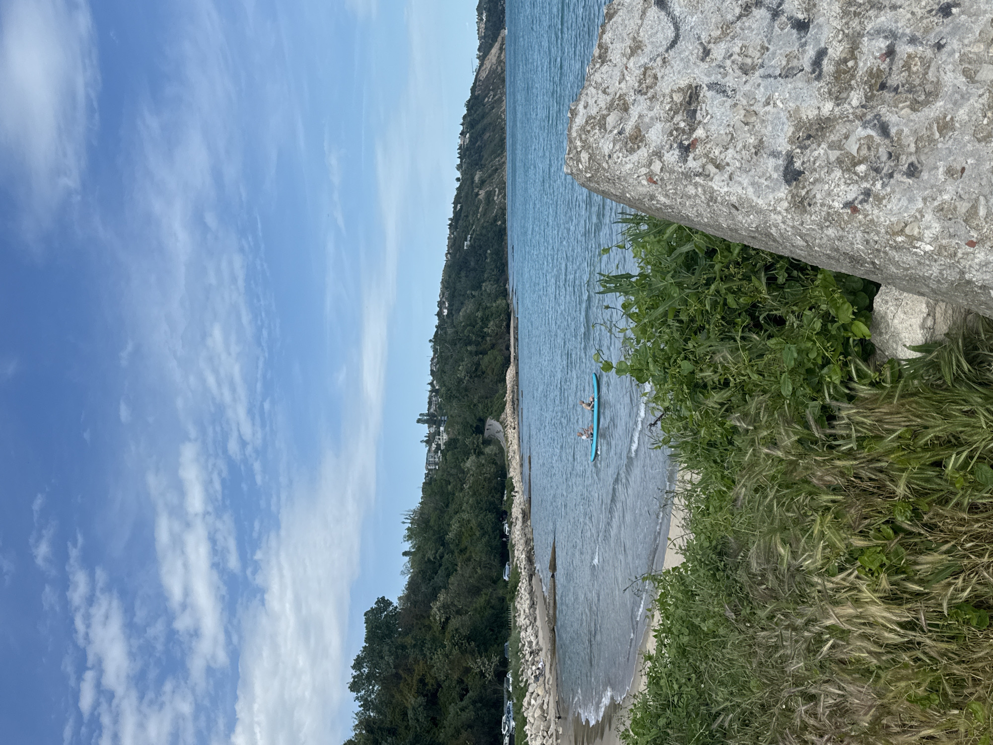 The image depicts a coastal scene near Varna, Bulgaria. The view is from a slightly elevated position, possibly a cliff or hillside, overlooking the sea and a sandy beach. A large concrete block is visible in the foreground, partially obscuring the view. Lush green vegetation, including grass and shrubs, grows around the concrete block.
In the distance, a person is paddling a blue stand-up paddleboard in the shallow water close to the shore. The shoreline is lined with trees and buildings, indicating urban development in the area.
The sky is blue with scattered white clouds, suggesting a clear day. The sea is calm, with gentle waves lapping on the shore. The overall scene captures a serene coastal environment.