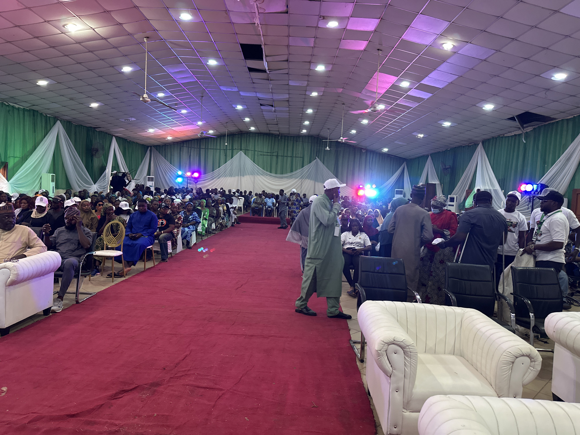 The image shows a large gathering of people in what appears to be an event hall located in an unknown city. The hall is adorned with green and white draped fabric along the walls. A red carpet stretches from the front to the back of the room, creating an aisle. Rows of chairs are placed on either side of the carpet, where people are seated. There are also white upholstered armchairs in the foreground. Many people in the hall are wearing white hats. The hall is lit by a combination of overhead lights and colorful spotlights. There are some visible holes in the ceiling. Some people are standing and interacting near the armchairs, while others are seated. The general atmosphere seems lively and event-oriented.