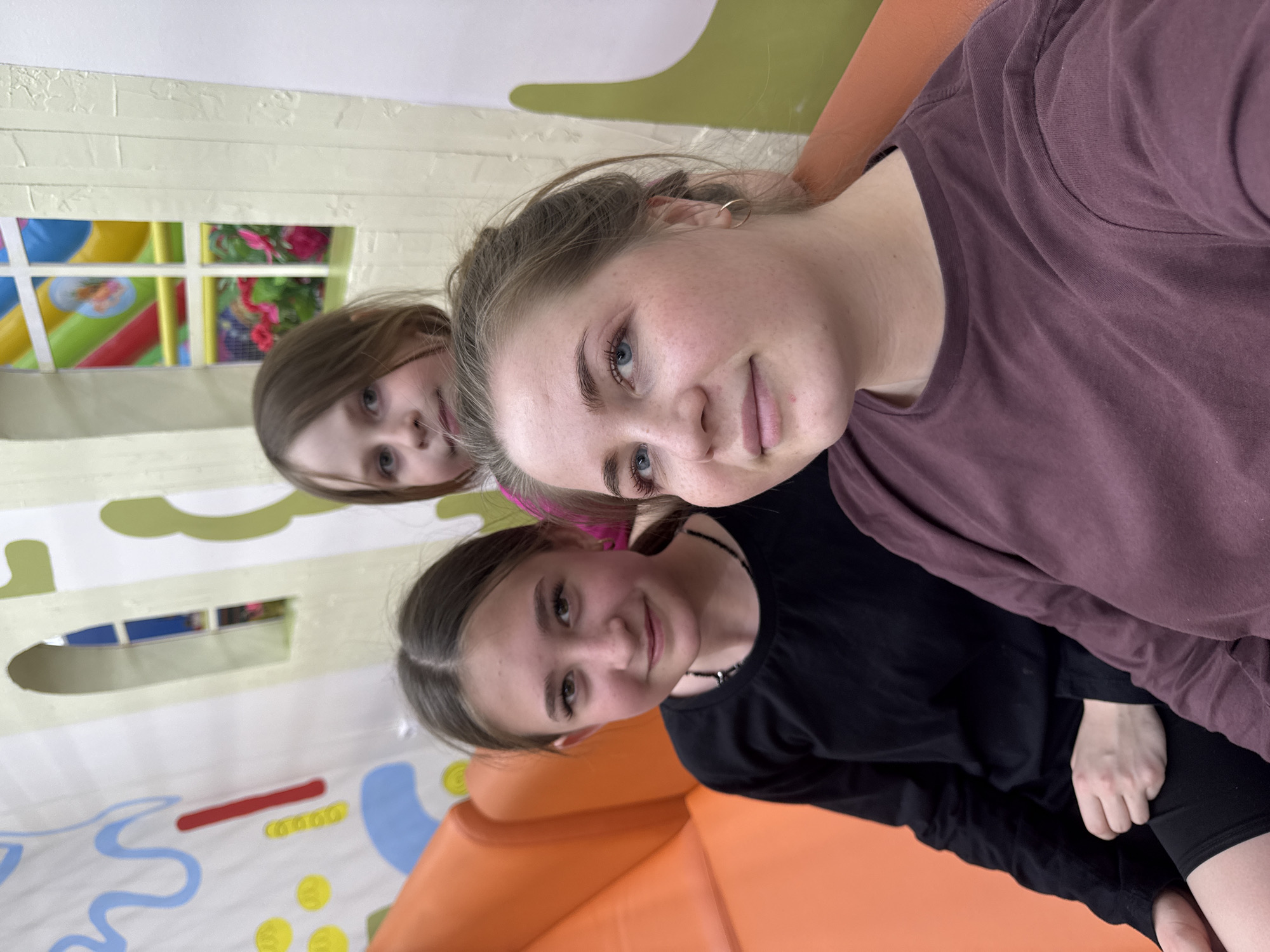 Three girls are posing for a selfie indoors in what appears to be an indoor playground or play area.
The girl in the upper right is wearing a mauve-colored long-sleeved shirt, her hair is pulled back, and she is looking at the camera. Below and to the left of her is another girl wearing a black long-sleeved shirt and a necklace. She is also looking at the camera. In the background, behind the second girl, is the face of a younger girl with brown hair.
The background features elements typical of a children's play area, including painted walls with rounded shapes and bright colors. There appears to be a playhouse with a window behind the youngest girl. An orange surface is visible at the bottom of the image.
It is not possible to confirm the location in Chaikovsky, Russia with certainty from the image.