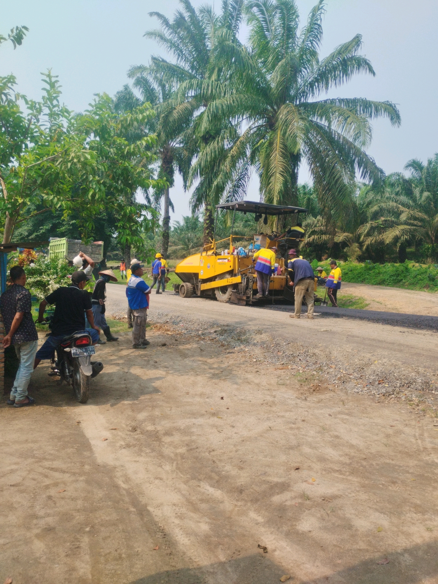 In Pagarbosi, Indonesia, a road construction project is underway amidst a backdrop of lush palm trees. A bright yellow asphalt paving machine, operated by at least two individuals wearing high-visibility vests, is actively laying down a fresh layer of asphalt on a dirt road. Several other workers, some also in safety vests, are present, observing the operation and likely involved in ancillary tasks. One person, wearing a conical hat, stands near the machine. A motorbike is parked on the side of the road, near a group of bystanders. The environment is rural, characterized by the dense vegetation of the palm plantation and a clear, bright sky. The overall scene depicts a busy work environment focused on infrastructure development in a tropical setting.