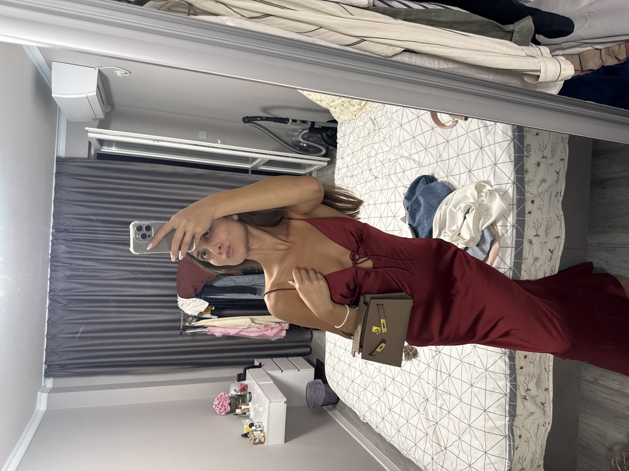 A young woman is taking a selfie in a bedroom in Burgas, Bulgaria. She is wearing a red satin slip dress and holding a small brown handbag. The room is messy, with clothes strewn on the bed and a clothing rack in the background. The woman is standing in front of a large mirror, and a vacuum cleaner is visible in the reflection.