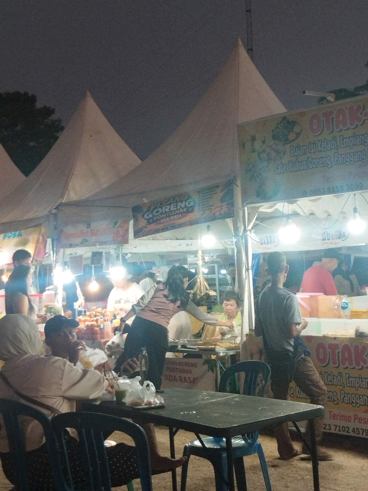 At night, an outdoor food stall area is depicted, likely a market or street food festival, in Parit Padang, Indonesia. The scene is dimly lit by artificial lights. In the foreground, a dark metal table with blue plastic chairs is set up, with a few people seated and interacting. One person is wearing a hijab and appears to be eating, while another, a male wearing a black cap, is drinking from a plastic bag.
In the background, several food stalls are visible, marked by canvas canopies. Signs for various food items like "Otak-Otak", "Pisang Goreng Pontianak", and "Sate Kukus/Goreng, Panggang" are displayed. People are seen ordering, preparing, and consuming food. There are also various food items and ingredients displayed at the stalls, including fruits and what appear to be snacks. The overall atmosphere suggests a busy and lively evening market.