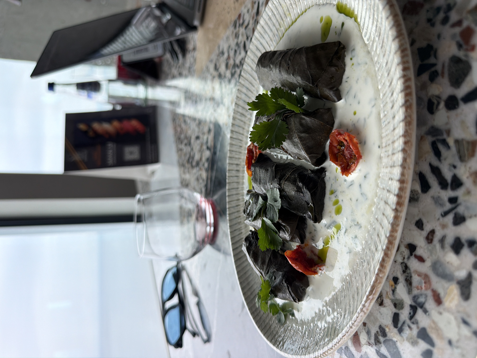 The scene depicts a plate of dolma (stuffed grape leaves) served in a creamy sauce and garnished with parsley and sun-dried tomatoes. The dish is presented on a decorative white plate. A glass of red wine and a pair of sunglasses are also present. A laptop and a bottle are visible in the background, along with a menu. The location is likely a restaurant or cafe in Batumi, Georgia. The table has a patterned terrazzo surface. The photo suggests a meal setting in a modern establishment.