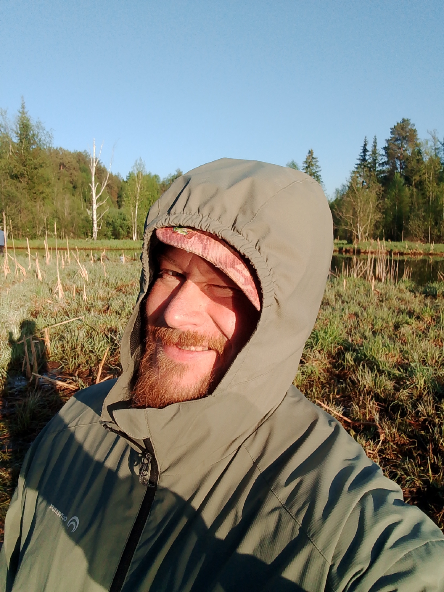 A man with a reddish-brown beard and wearing a light green hooded jacket stands in a field of tall grass near a body of water. The man is smiling slightly and appears to be taking a selfie. Behind him is a marshy area, and beyond that, a forest. The sky is blue and mostly clear. The location is Chaikovsky, Russia.
