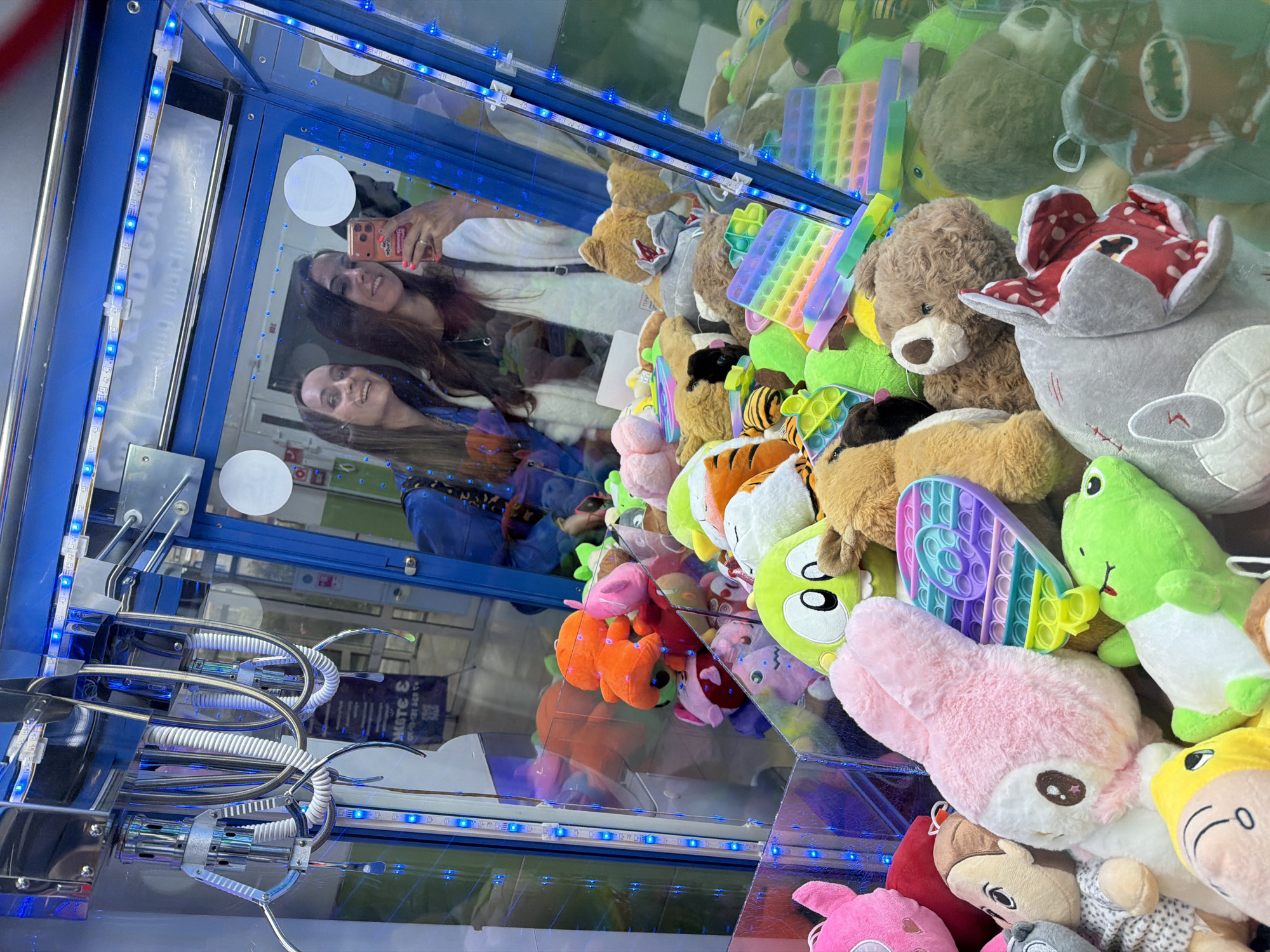 The image shows the interior of a claw machine, stocked with plush toys and pop-it fidget toys. The machine is illuminated with blue LED lighting. The claw mechanism is visible in the foreground. Reflected in the glass of the machine are two women taking a selfie. It is impossible to determine if the location is in Birobidzhan, Russia, based on visual information.