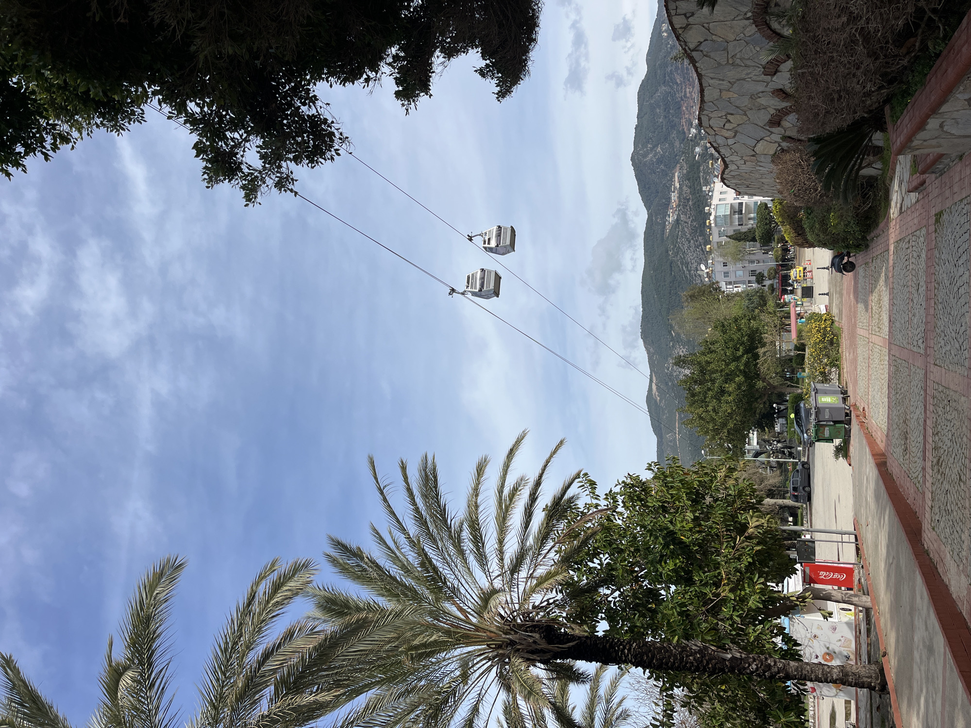 The image depicts a view in Alanya, Turkey, showing two cable cars suspended on wires against a partly cloudy sky, framed by foliage. Below, a palm tree and other greenery are visible. In the distance, a mountain slope is dotted with buildings. Adjacent to the lower right, a stone wall borders a paved walkway. A Coca-Cola vending machine is also seen on the bottom right.