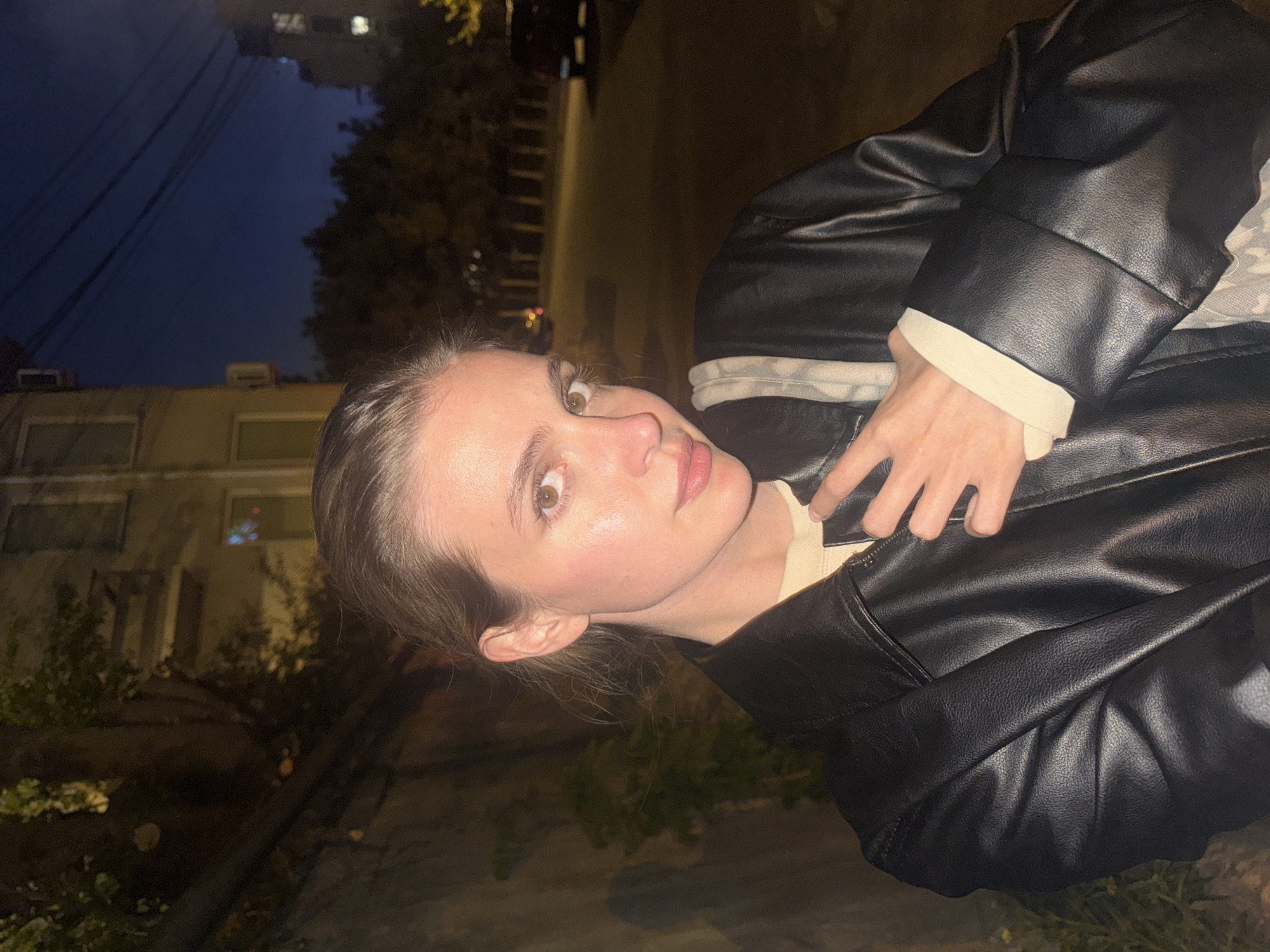 A young, fair-skinned woman with light brown hair is positioned in the foreground of the image. She is wearing a black leather jacket layered over a light-colored top. The focus is on her face, which is directed toward the viewer.
The background reveals an urban environment. Buildings can be seen, and streetlights illuminate the scene, suggesting it is nighttime. Trees line the street. The presence of architecture and street lighting indicates this is likely a city setting. Based on the information provided, it is possible this image was taken in Tbilisi, Georgia, however, without specific landmarks, verification is impossible.