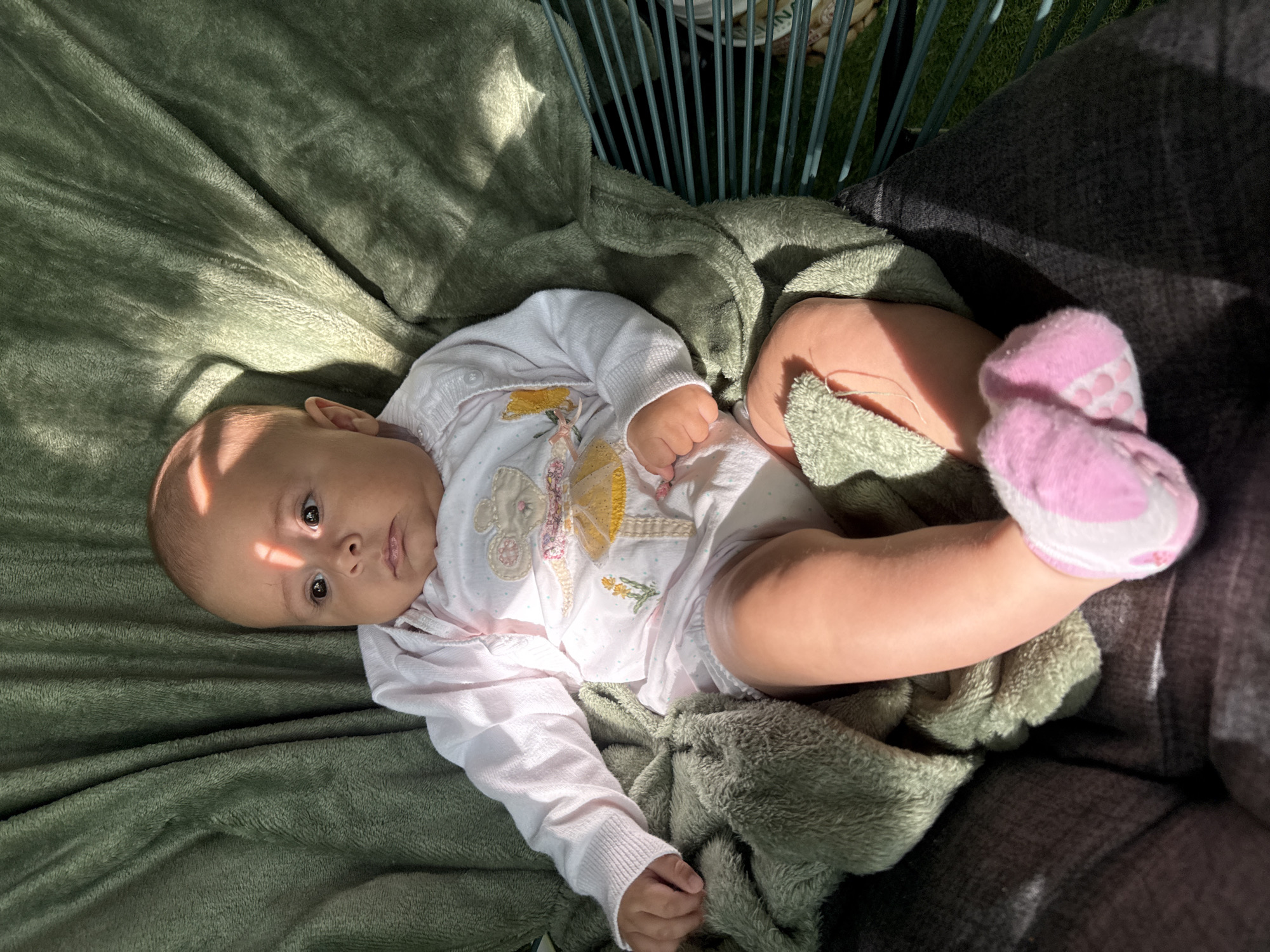 A baby is nestled in a green hammock or chair outdoors, covered by a soft green blanket. Sunlight streams down, casting striped shadows across the baby's face and body. The baby is wearing a white outfit with a pink trim and a design of a mouse in a tutu. Pink socks with white accents are on the baby's feet. The hammock or chair appears to be made of dark gray fabric and green metal bars, suggesting it might be located on a patio or garden area. The background shows hints of greenery and possibly a lawn. The overall scene evokes a sense of peacefulness and relaxation. The location is stated to be in the city of Plovdiv, Bulgaria.