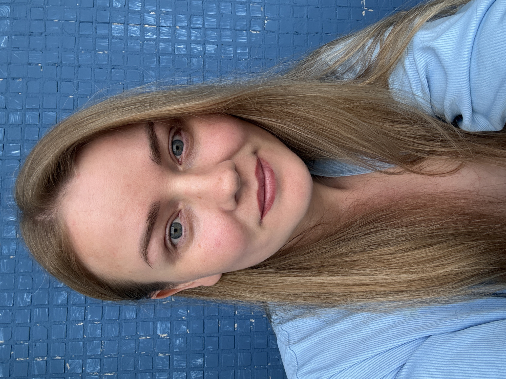 A close-up selfie of a young woman with long blonde hair is presented against a textured blue background. The woman is facing the camera, and her head is slightly tilted to her right. She is wearing a light blue collared shirt. Her hair is parted in the middle and falls down on either side of her face. Her eyes are blue-green, and she has fair skin with rosy cheeks. Her lips are slightly parted, and she is wearing a muted berry-colored lipstick. The blue background appears to be made of small, rectangular tiles. The image does not provide any information or context about the city of Chaikovsky, Russia.