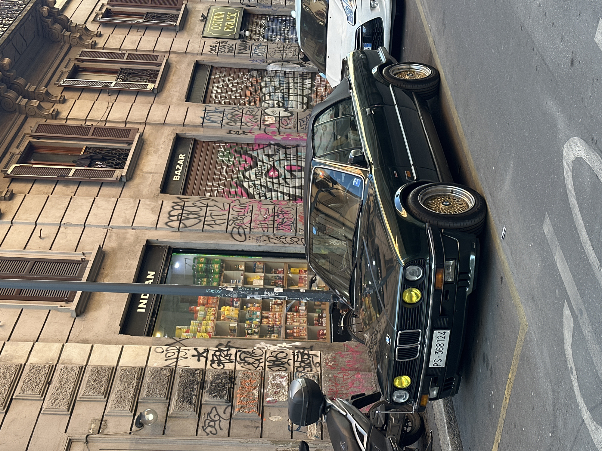 The image shows a street scene in Milan, Italy, with an old dark-green BMW convertible parked next to a scooter along the curb. The BMW has a license plate that reads "PS-368124". The background features an old building with several storefronts covered in graffiti, including a shop named "INDIAN" with groceries visible inside, and a partially visible storefront marked "BAZAR". Above these, other shops are shut with metal shutters also covered in graffiti, including "OSTERIA PUGLIESE" with "IDEO SODA ZI RODRIO" painted beside it. The building's facade is classic, with multiple windows and decorative architectural details. The street shows signs of wear, with markings and visible lines. The image is taken from a slightly rotated perspective.