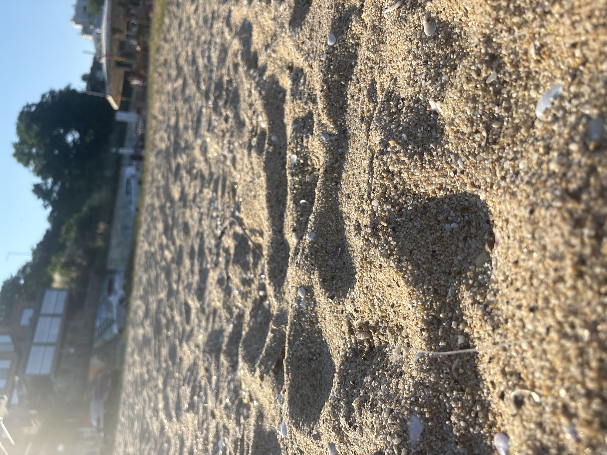 The image shows a close-up view of sand on a beach. The sand is light brown and appears to be made up of small grains and pebbles. There are several footprints impressed in the sand, suggesting recent activity. In the background, out of focus, there are hints of a building with windows, trees, and a clear blue sky, indicating an outdoor setting. The lighting suggests it might be morning or late afternoon, with shadows cast by the footprints. There is no visible text in the image. The overall impression is a serene beach scene with signs of human presence.
